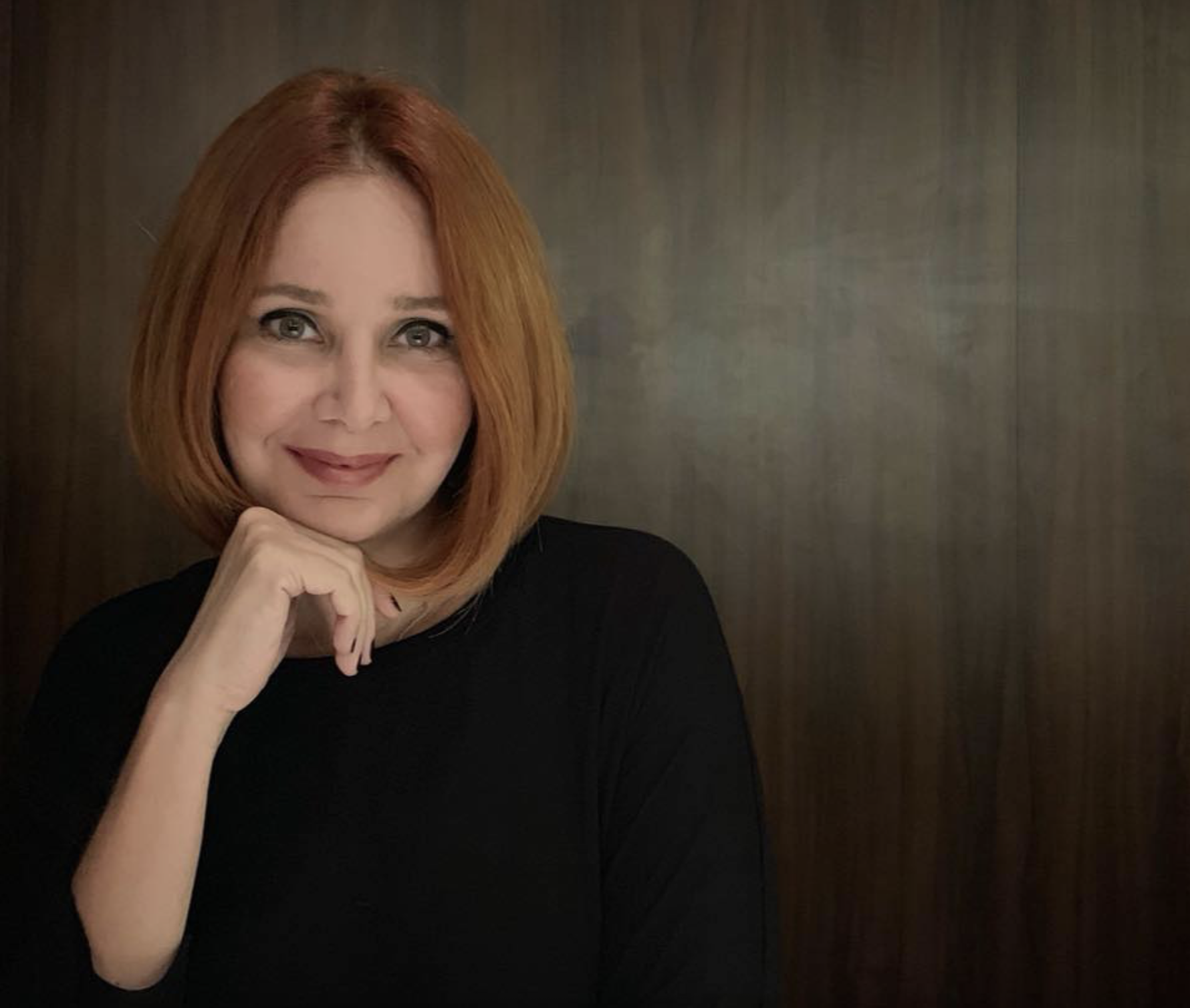 A woman with shoulder-length red hair, wearing a black top, is smiling and resting her chin on her hand, in front of a dark wooden wall.