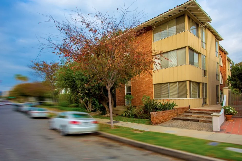 A multi-story apartment building with a brick and yellow exterior, surrounded by plants and trees, adjacent to a sidewalk and parking lot with several blurred cars, under a partly cloudy sky.