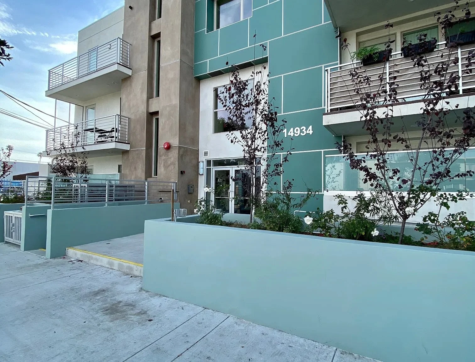 Modern apartment building with balconies, green and beige exterior, and the address 14934 visible on the side. There are small trees in front, and the sidewalk has a gentle slope.