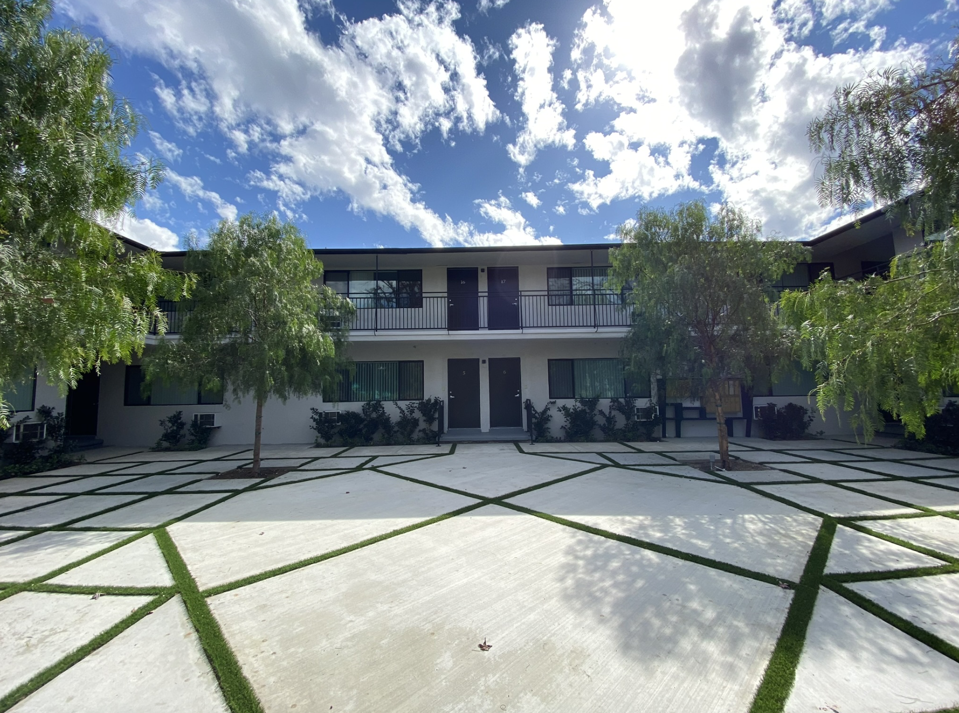 Exterior view of modern two-story apartment building with trees, a paved courtyard, and a partly cloudy sky.