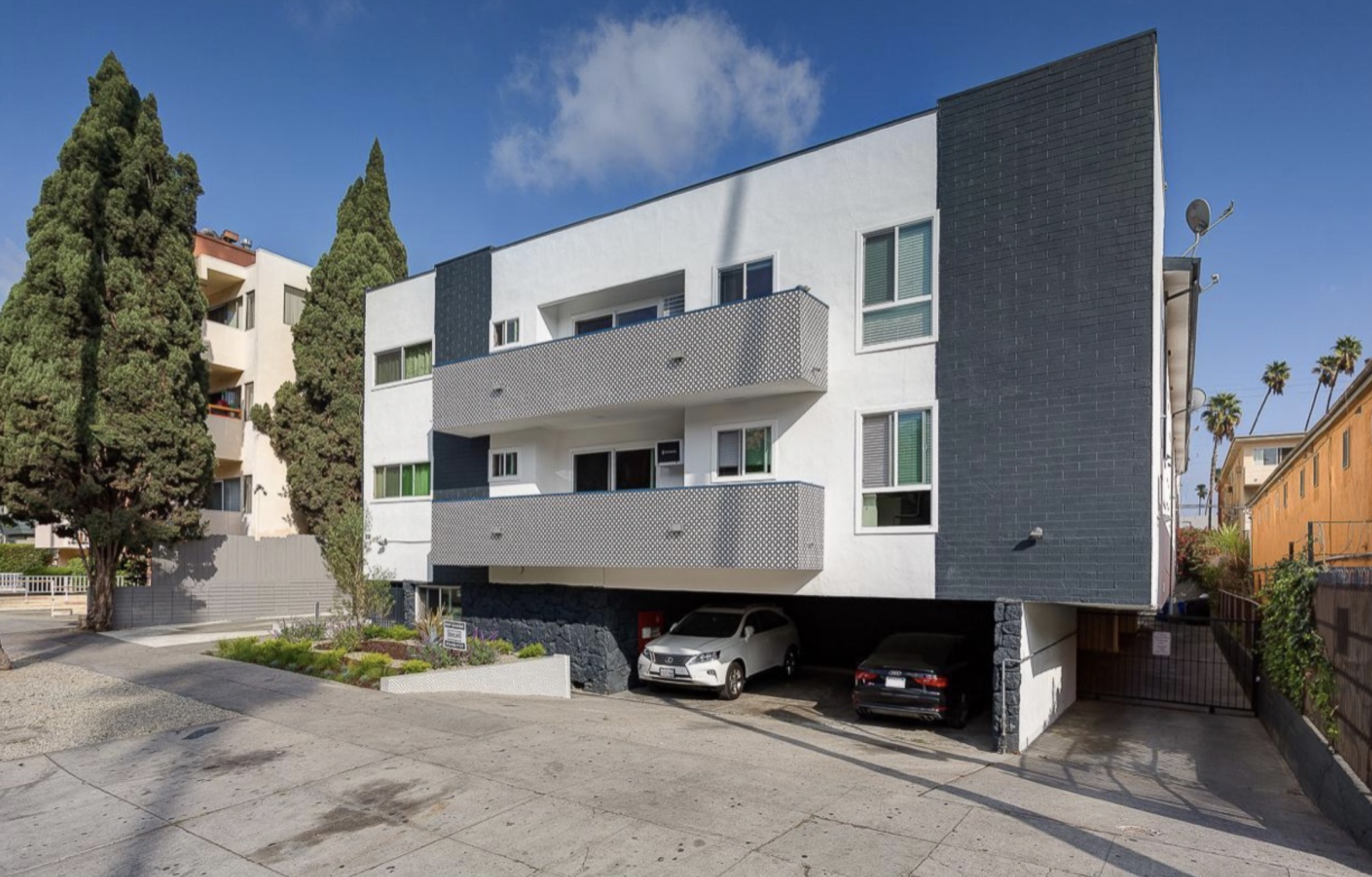 A modern four-story apartment building with white and dark gray exterior walls, balconies, and large windows. There are two cars parked under the building and tall trees in the background, with some palm trees visible to the right.
