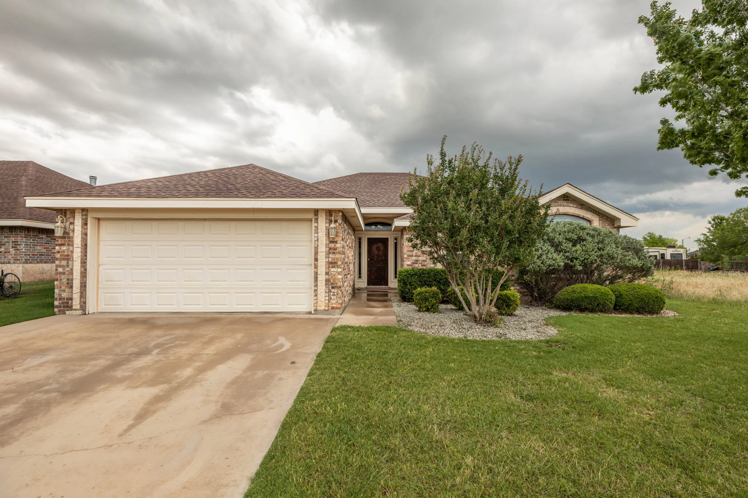 Front view of a suburban house with a brown shingle roof, brick exterior walls, a white garage door, a dark front door, and neatly landscaped yard with trees and bushes under a cloudy sky.