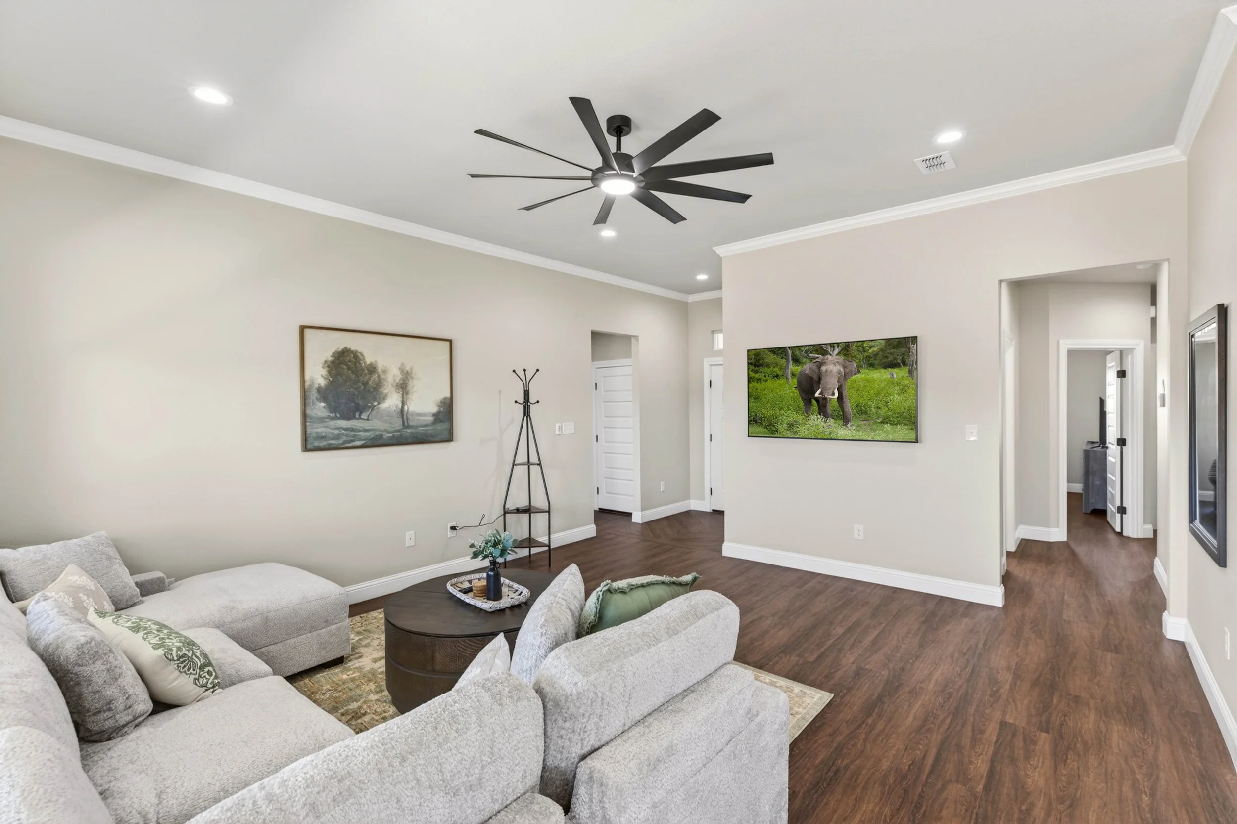 Living room with beige walls, a large sectional sofa, a round coffee table, wall art, a coat rack, and a wall-mounted television displaying an elephant in a green field. Ceiling fan with multiple blades.