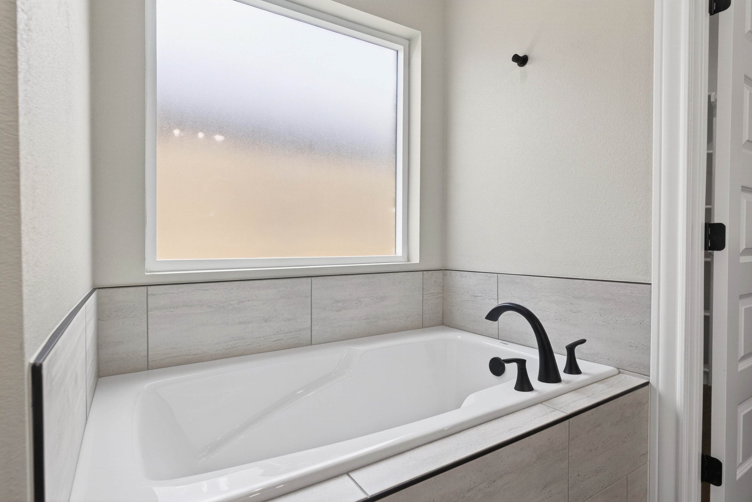 A white bathtub with black fixtures and a frosted window above it in a bathroom with beige tiled walls.