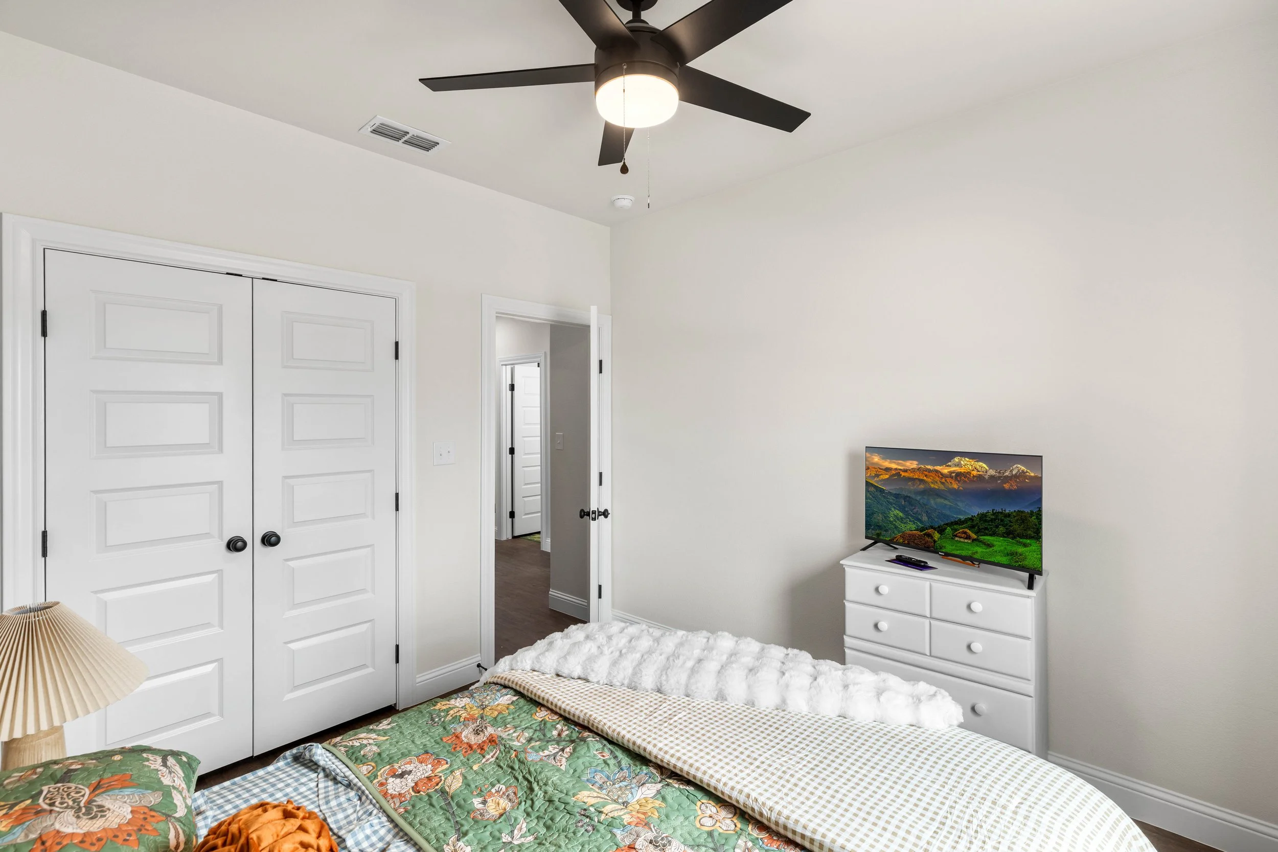 A bedroom with white walls, a ceiling fan, a doorway, and a television on a white dresser. The bed has a floral quilt and a furry white blanket.