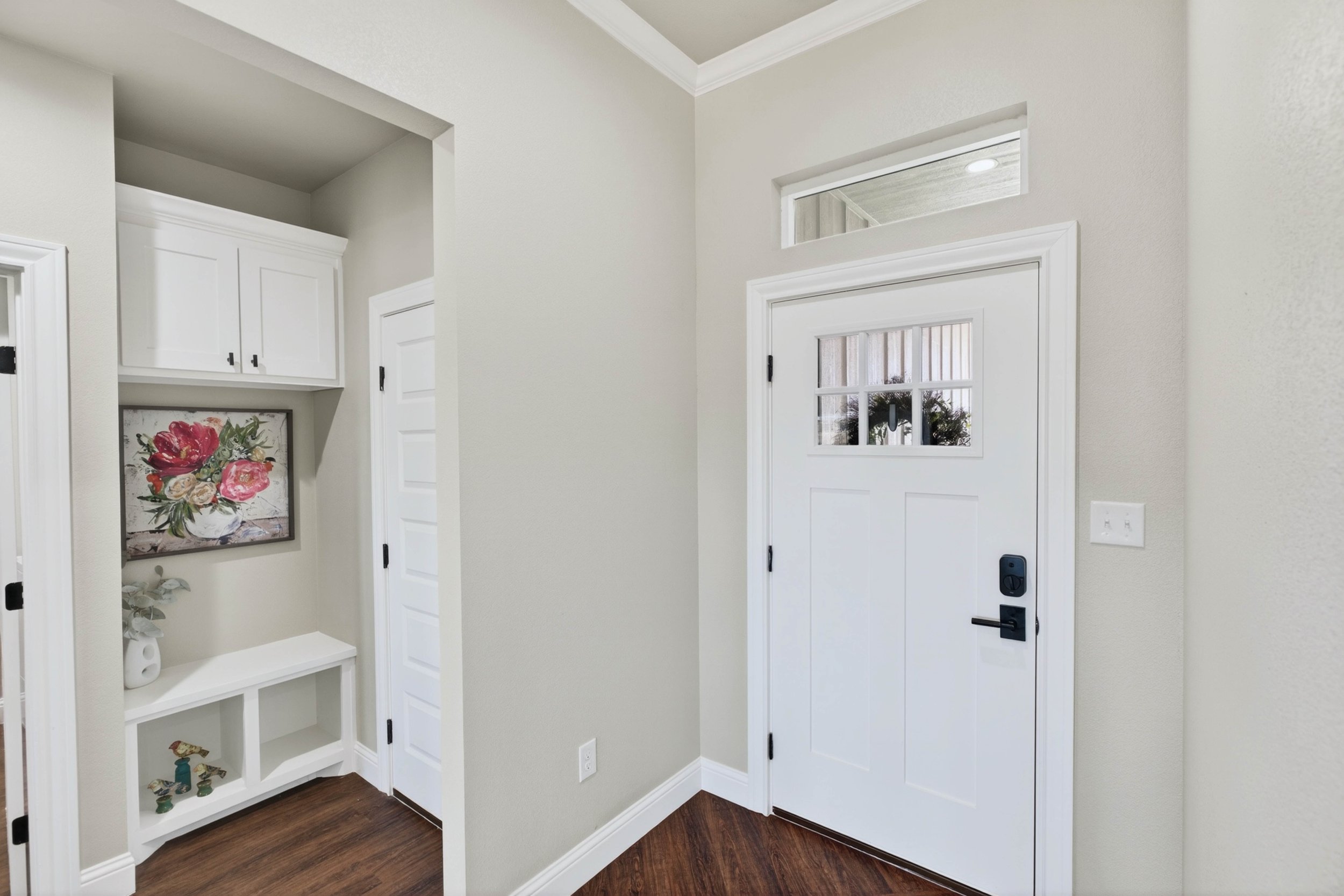 Interior view of an entryway with a white door and a small window above it, a built-in white shelf with decorative items, a framed floral artwork, and hardwood flooring.