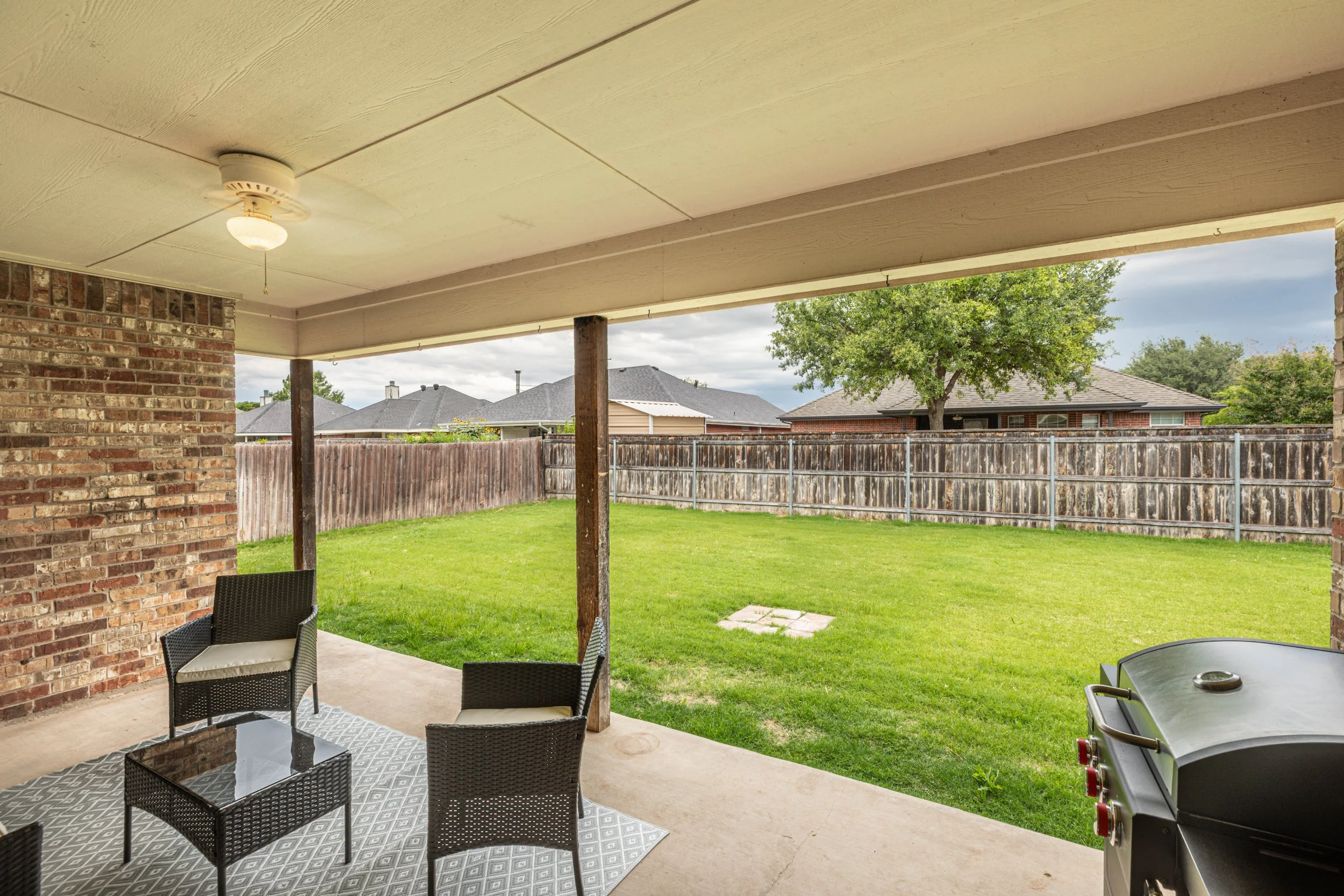 Backyard patio with outdoor furniture, a barbecue grill, and a view of a fenced lawn with trees and rooftops of neighboring houses.