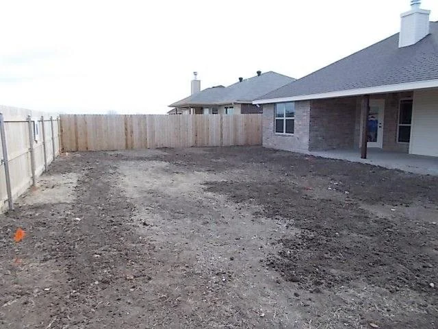 Empty backyard with dirt ground, surrounded by a wooden fence on the left and back, and a house with a covered patio on the right.