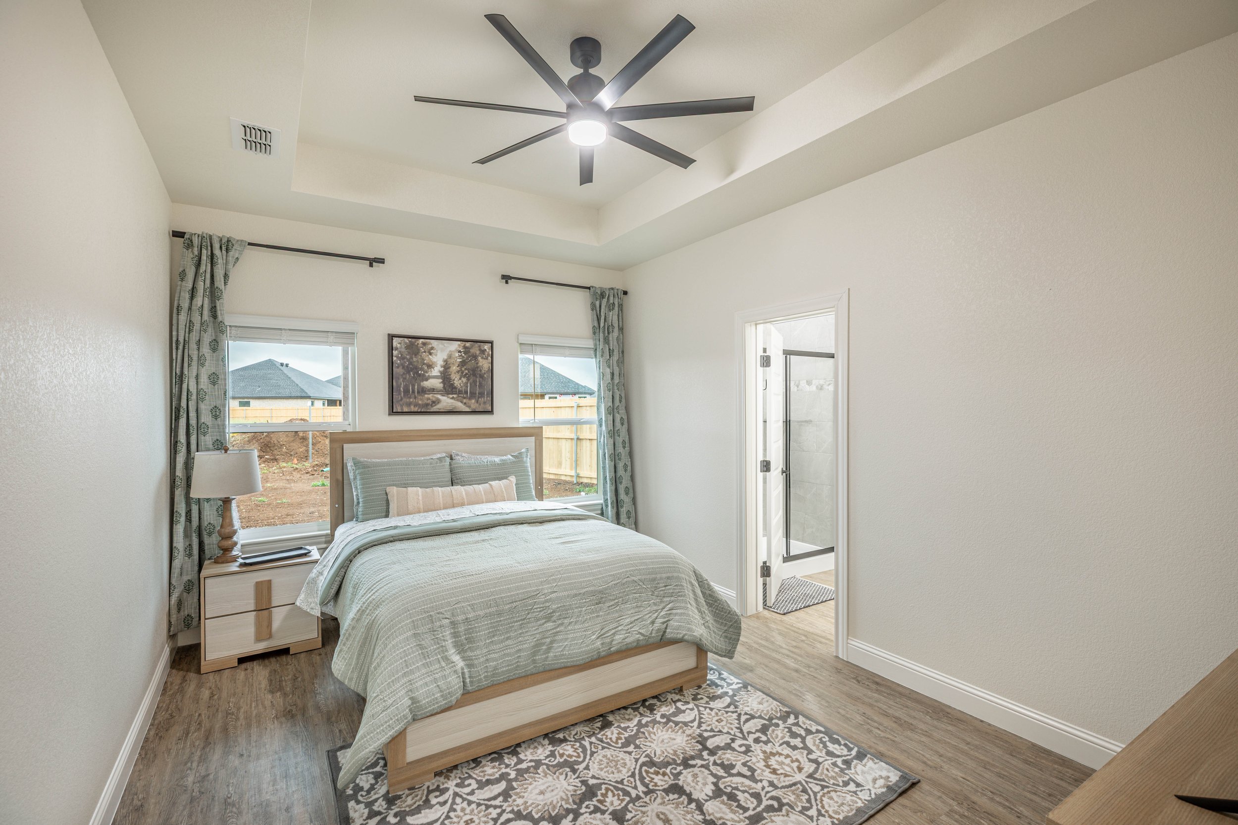 A bedroom with a wooden bed frame, green and beige bedding, a nightstand with a lamp, large windows with curtains, and a ceiling fan.