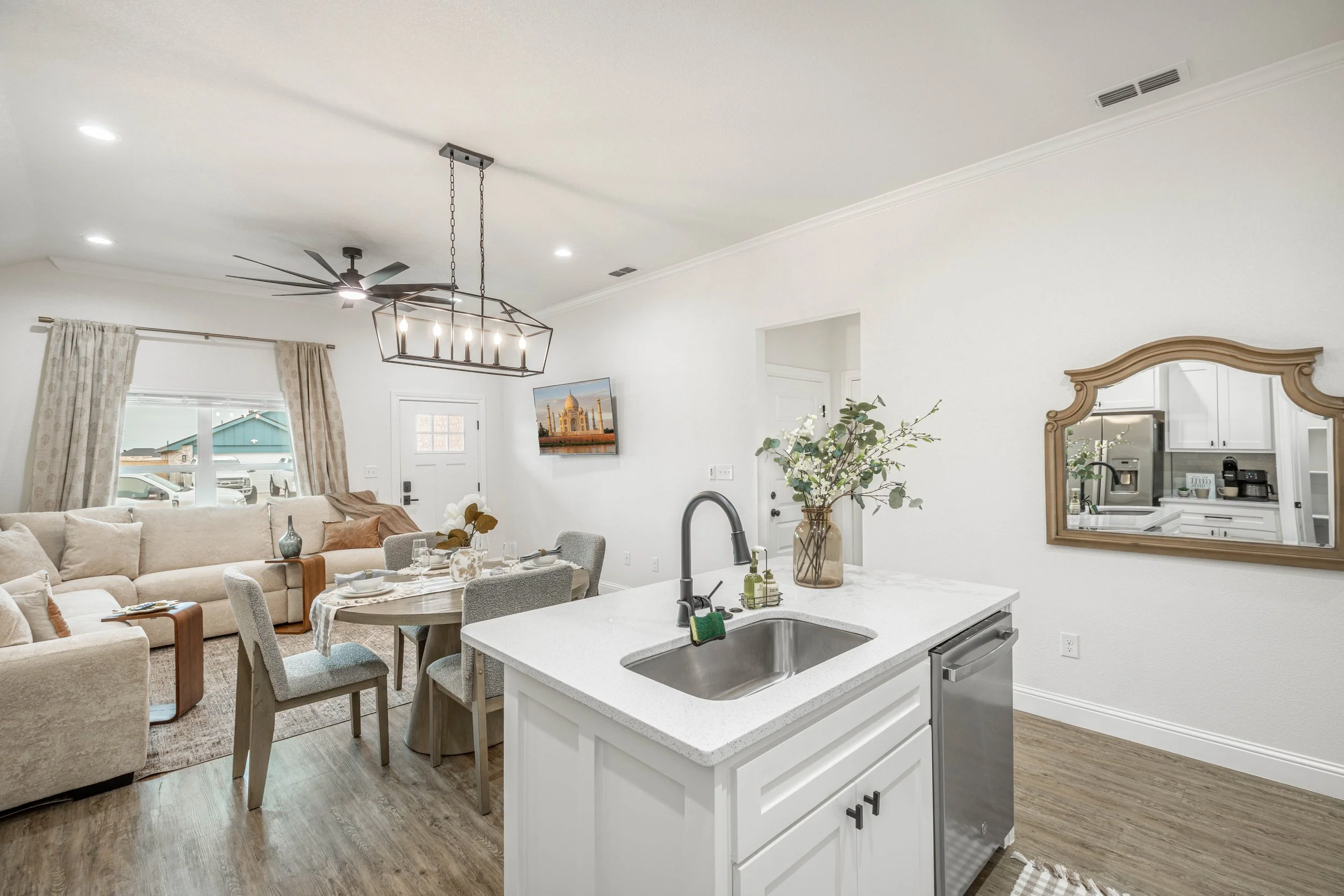 Open-concept living and dining area with a beige sectional sofa, dining table with chairs, and a kitchen island with sink; hanging light fixture, ceiling fan, and framed photo of Taj Mahal on the wall.