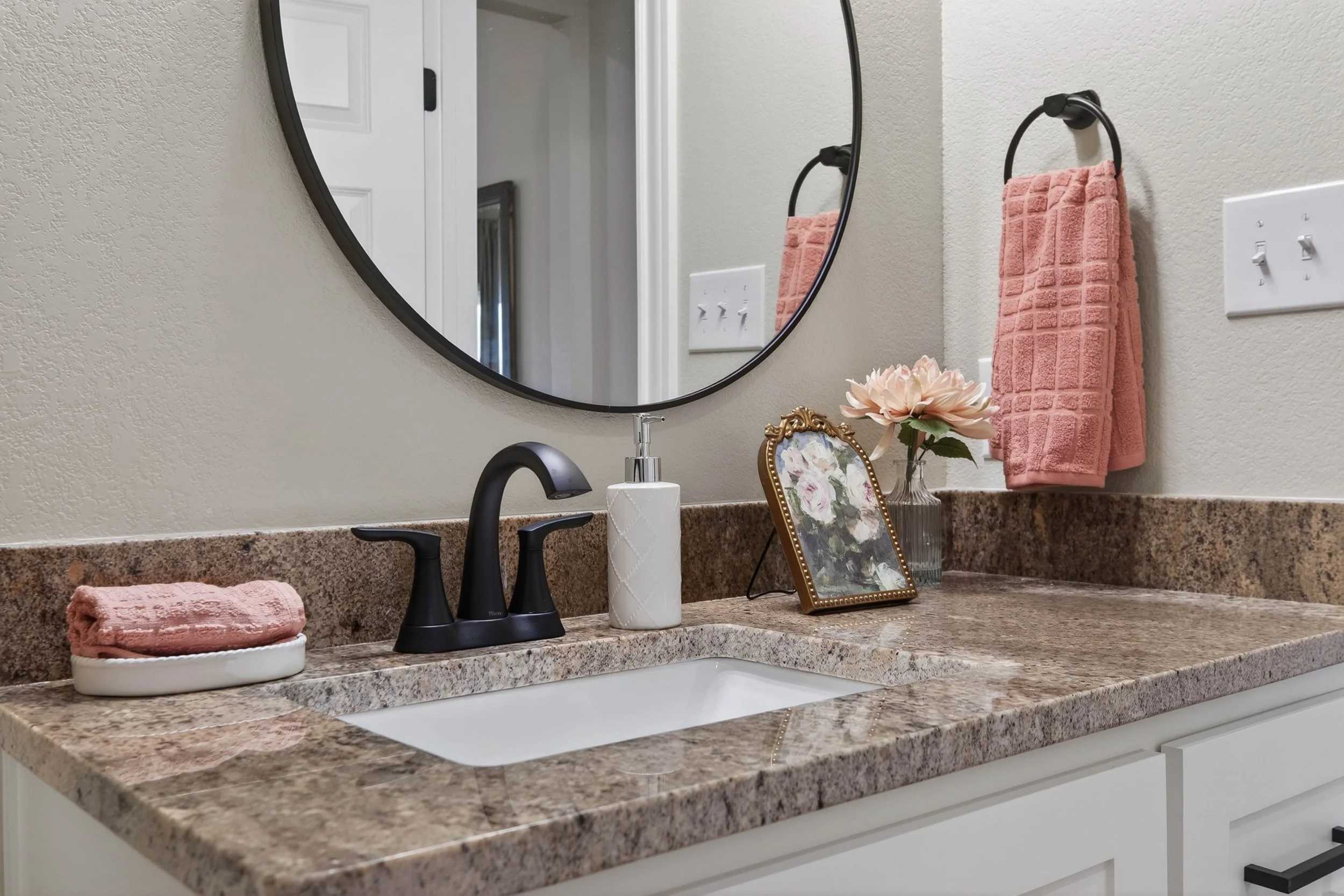 Bathroom countertop with a mirror, pink towel on a black ring holder, a small framed floral artwork, a soap dispenser, a floral towel, and a paper towel roll.