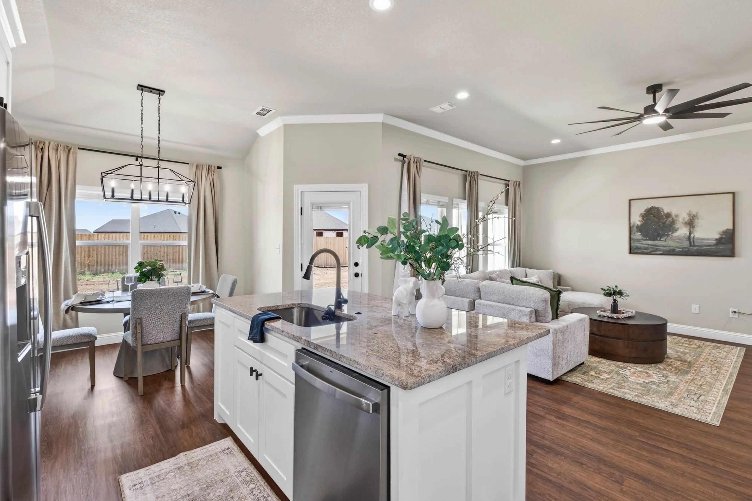 Open-concept living room and kitchen with beige walls, hardwood flooring, a white kitchen island with a granite countertop, a white sofa, a round coffee table, a large wall painting of trees, and a ceiling fan.