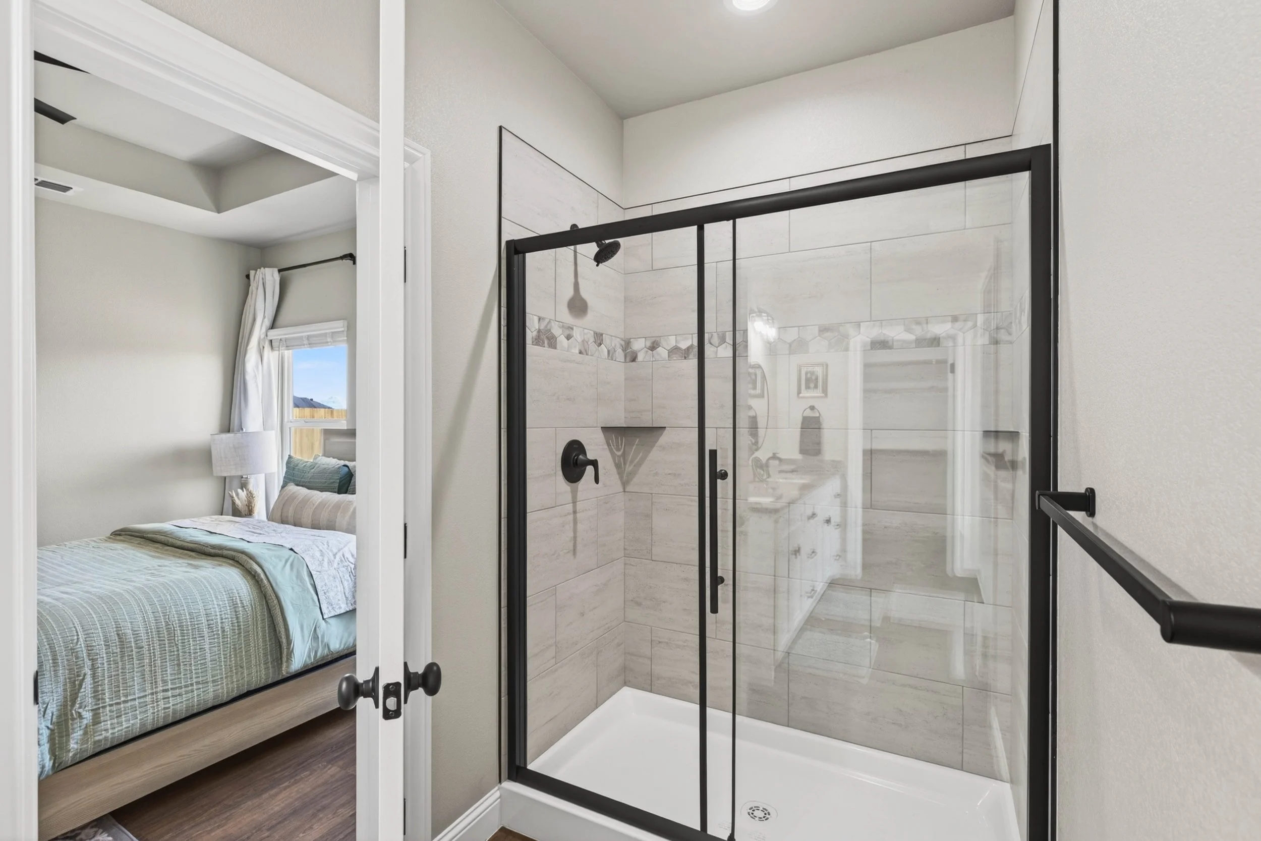 A modern bathroom with a glass shower enclosure featuring black framing and beige tile walls, adjacent to a bedroom with a bed and window with white curtains.