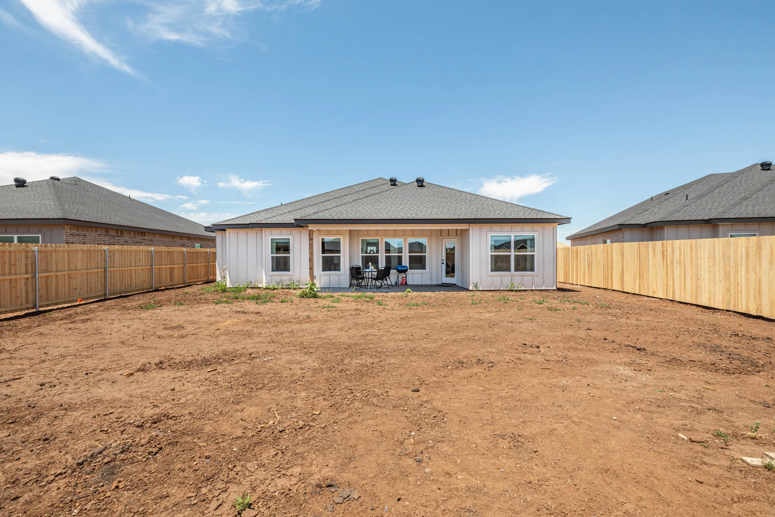 Backyard with dirt ground, wooden fence, and a white house with gray roof, patio furniture, barbecue grill, and sky with clouds.