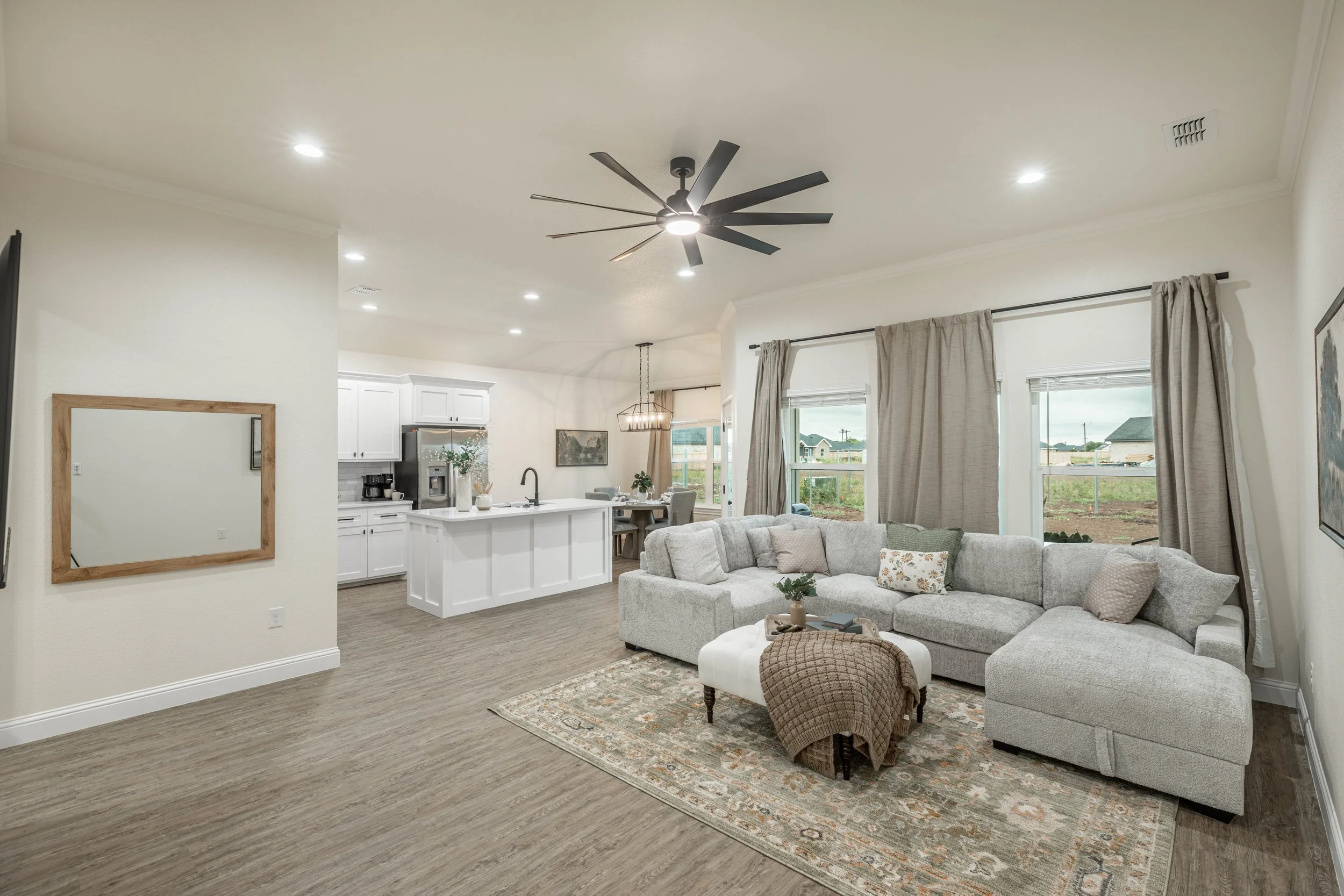 Open-concept living room and kitchen with beige walls, large windows, gray upholstered sofa, area rug, ceiling fan, and natural light.