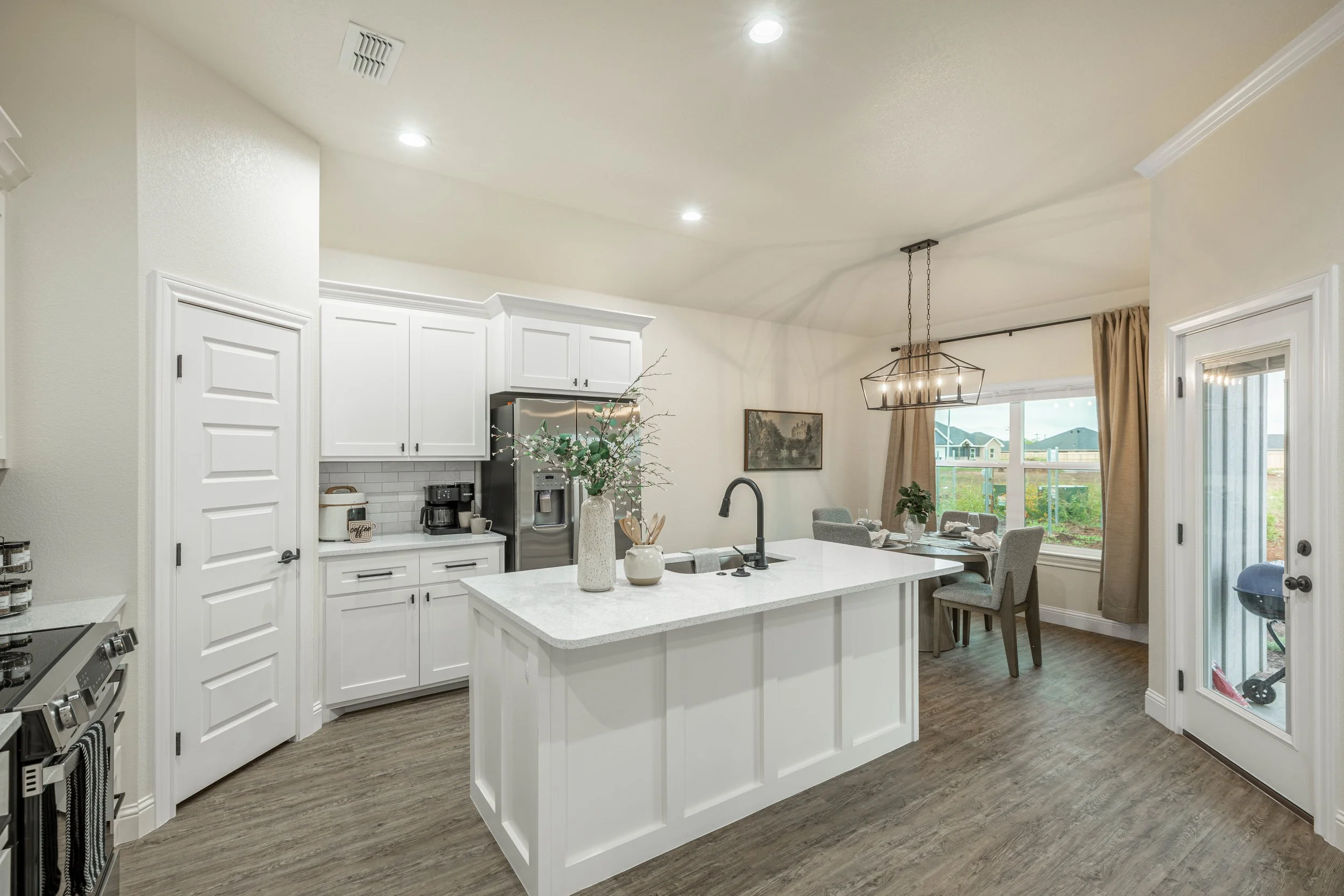 A bright, modern kitchen with white cabinets, a large white island, stainless steel refrigerator, and a dining area with a table and chairs near a window with beige curtains. There is a hanging chandelier and a view of a backyard outside.