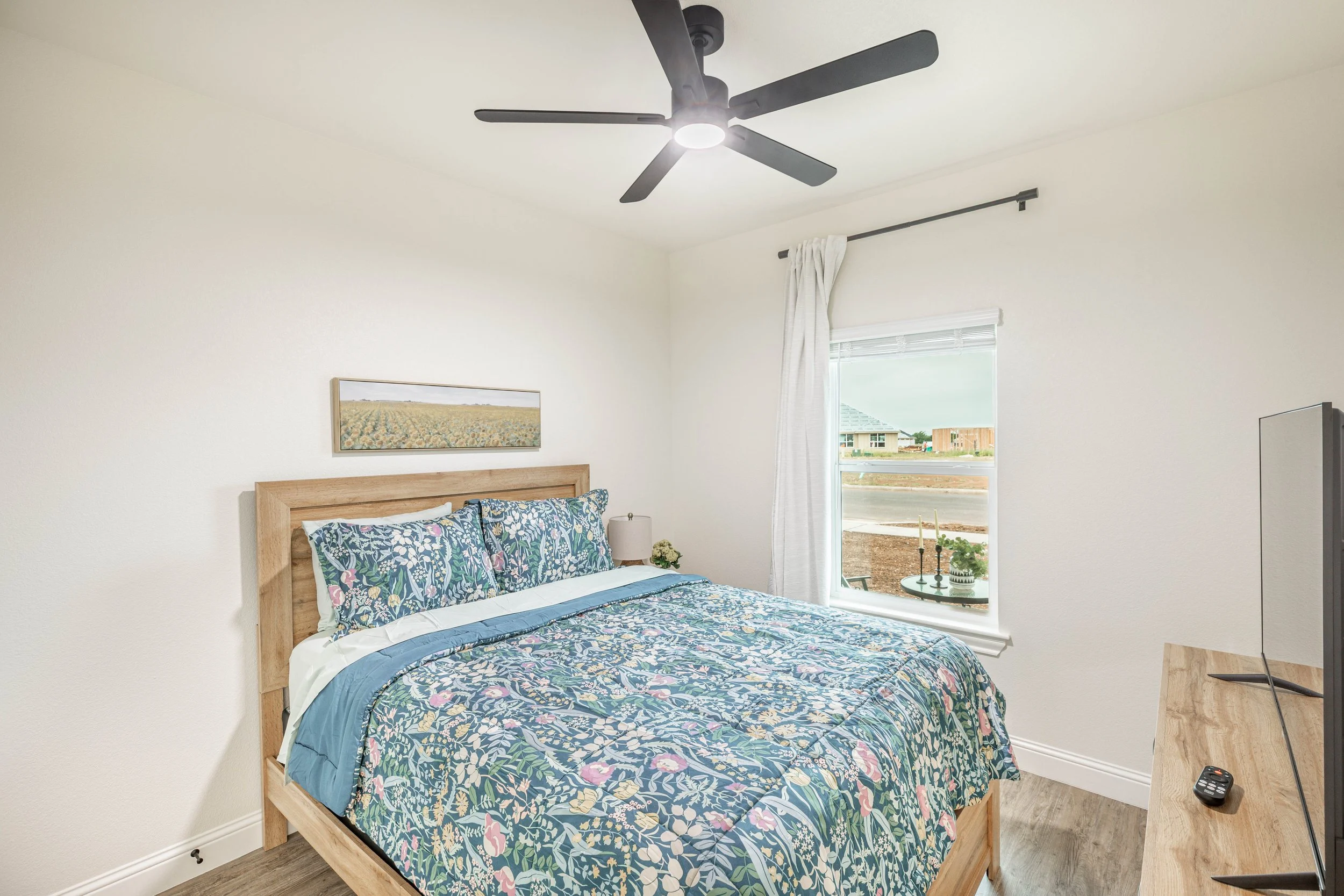 A bedroom with a wooden bed frame, floral bedding, and pillows. A painting hangs on the white wall above the bed. A window with white curtains shows a view of houses and a street outside. A ceiling fan with five black blades is on the ceiling, and a 