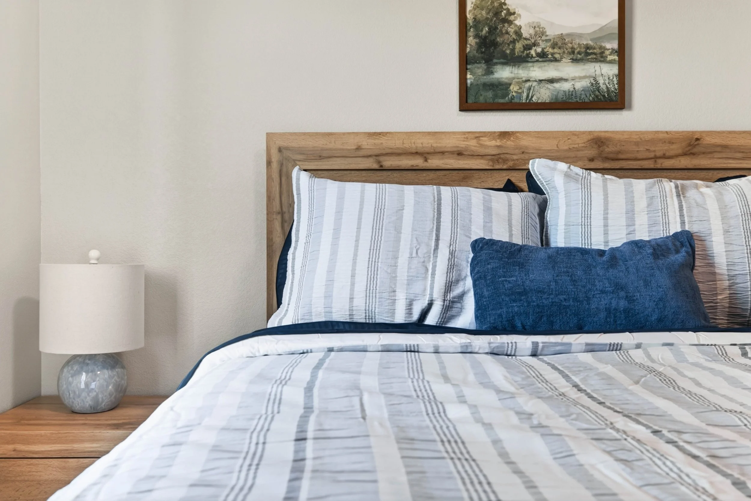 A neatly made bed with white and gray striped bedding, a dark blue pillow, and an oak headboard in a bedroom. A round white table lamp is on a wooden nightstand beside the bed. A landscape painting hangs on the wall above the headboard.