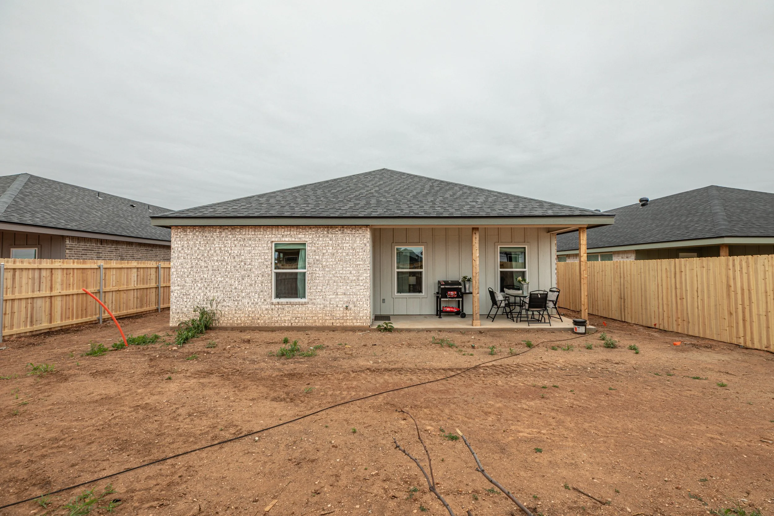 Backyard of a house under construction with exposed dirt, wooden fences, and a patio area with outdoor furniture and a grill.