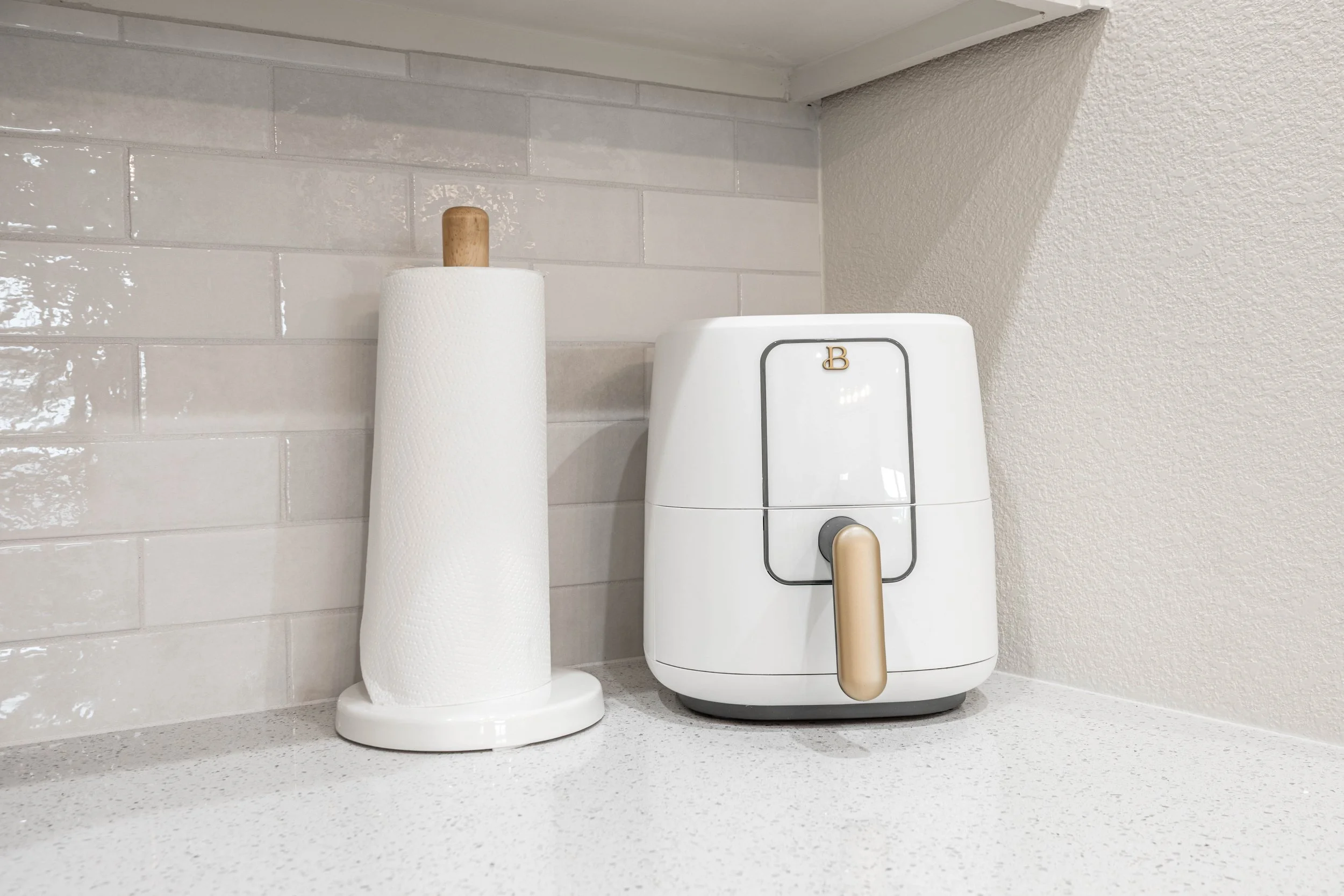 A paper towel holder with a roll of paper towels and a vintage-style white and gold bread box on a light-colored kitchen countertop, with a textured white tile backsplash and wall.
