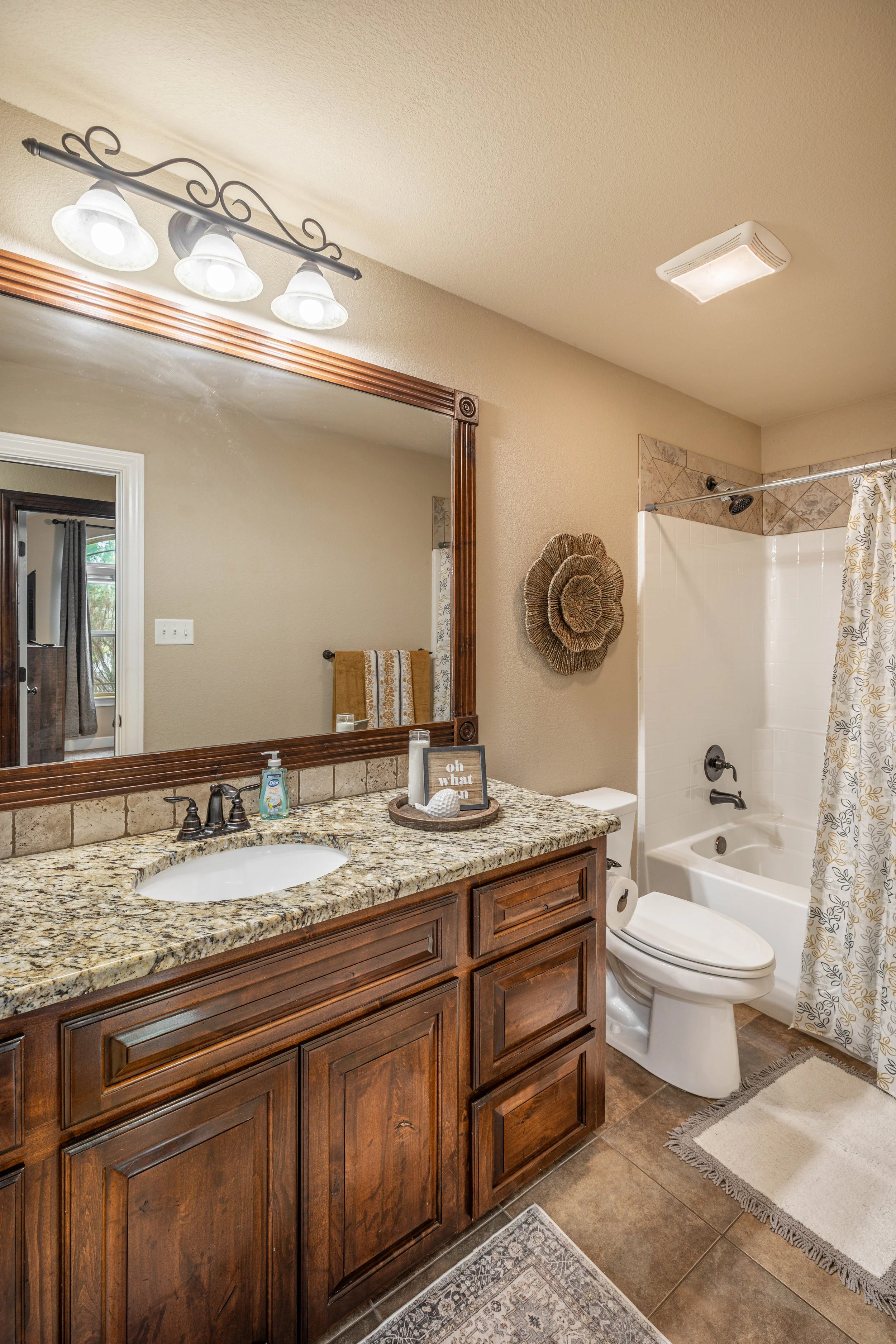 Bathroom with granite countertop vanity, large wall mirror, decorative wall art, toilet, bathtub with shower curtain, and ceiling lights.