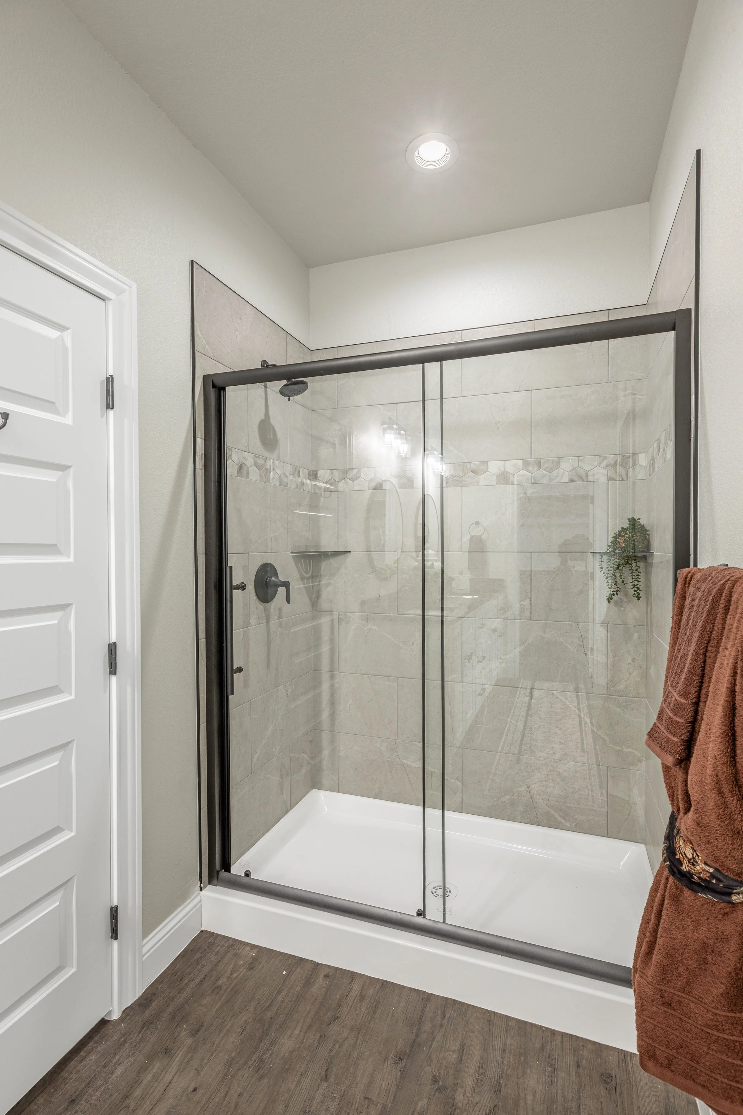Bathroom with a walk-in shower enclosed by glass with a metal frame, beige tiled walls, a showerhead, a control handle, a small green plant, a brown towel hanging on a hook, white ceiling with a recessed light, a white door, and wood flooring.