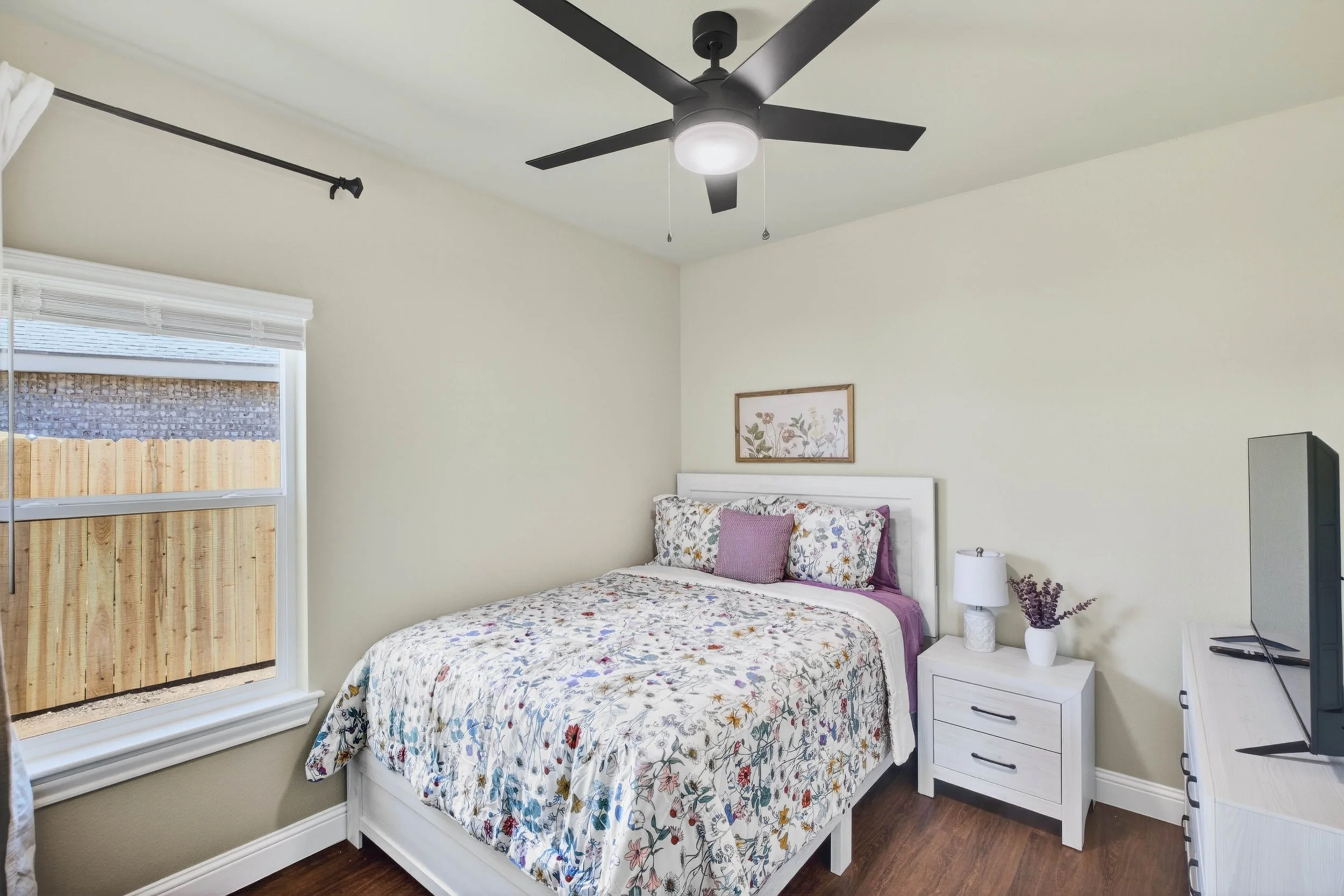 A bedroom with a white bed frame and floral bedding, a nightstand with a white lamp and vases, a window with white blinds, a framed floral print on the wall, a black ceiling fan with light, and a television on a white dresser.