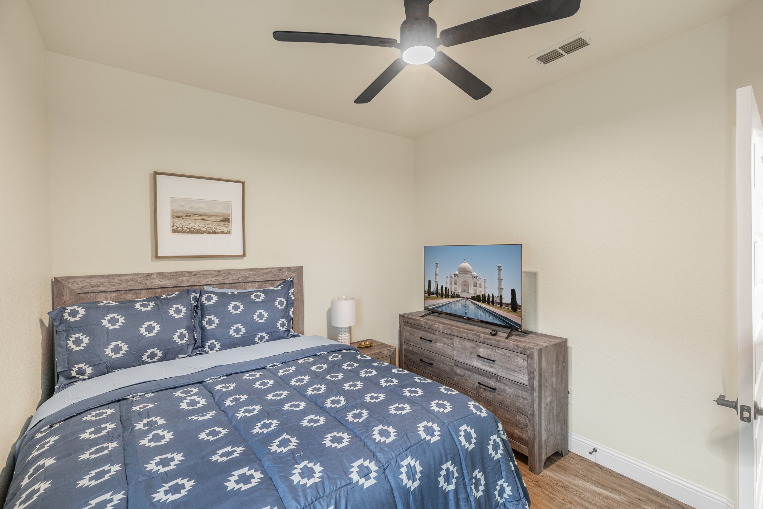 A bedroom with a wooden bed frame and matching dresser. The bed has blue and white patterned bedding. There is a framed picture on the wall above the bed, a white table lamp on the dresser, and a television displaying the Taj Mahal. A ceiling fan wit
