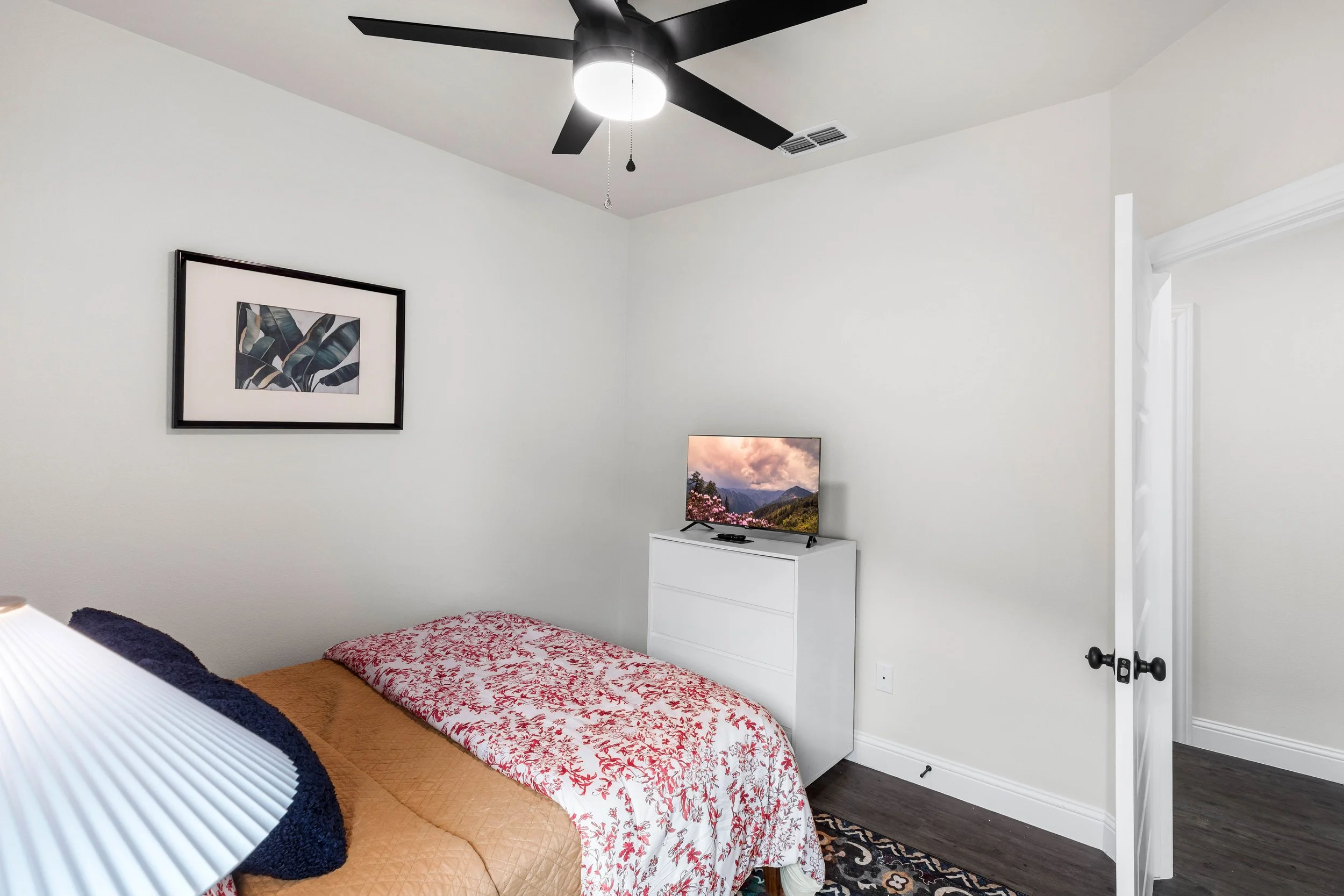A bedroom featuring a bed with a floral bedspread, a white dresser with a flat-screen TV on top, a picture of leaves framed on the wall, a ceiling fan with lights, and a portion of a doorway with white walls and crown molding.