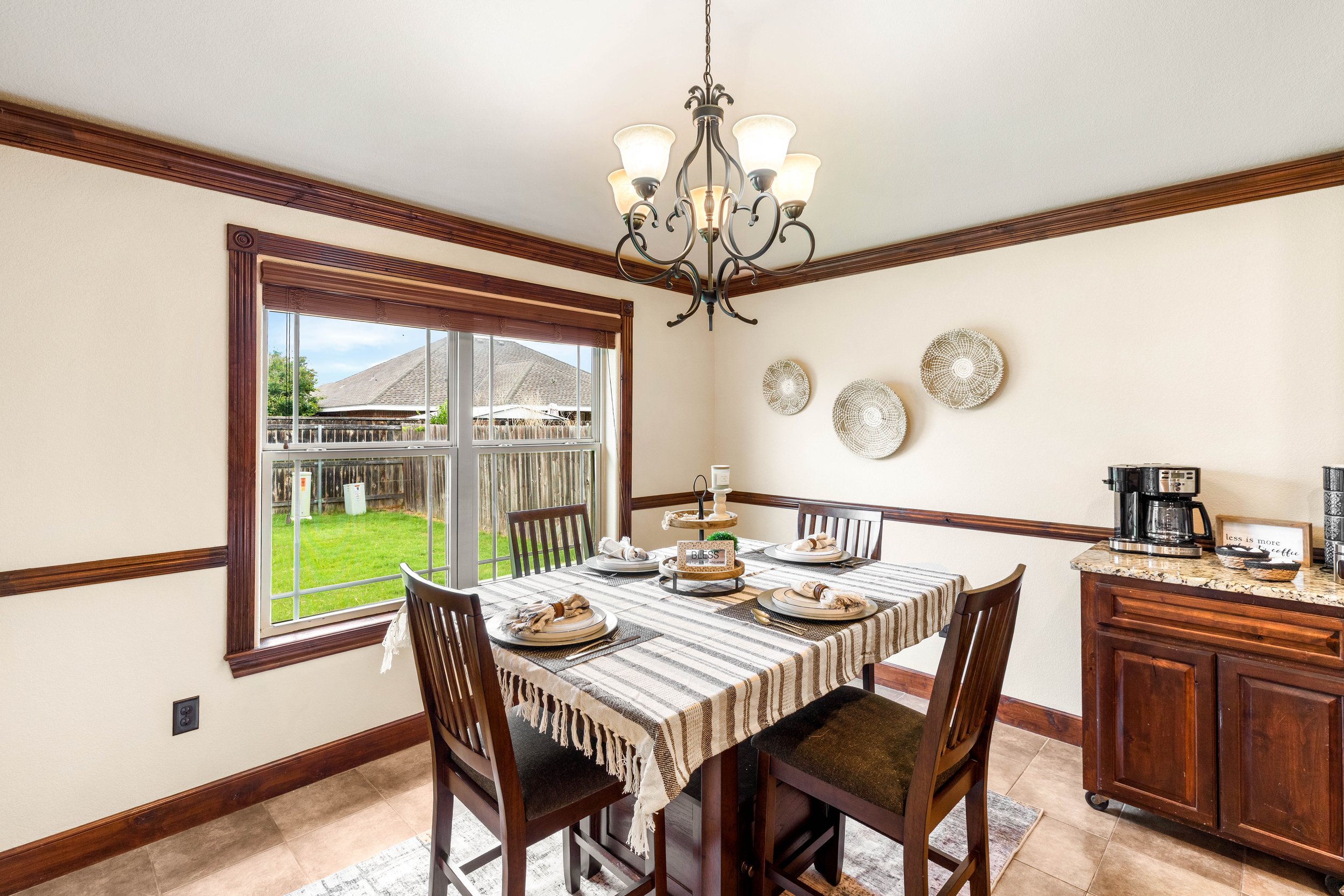 A dining room featuring a wooden table with a striped tablecloth, set with plates, napkins, and wine glasses. Six wooden chairs surround the table. There is a window with a view of a backyard with a wooden fence, grass, and neighboring houses. The ro