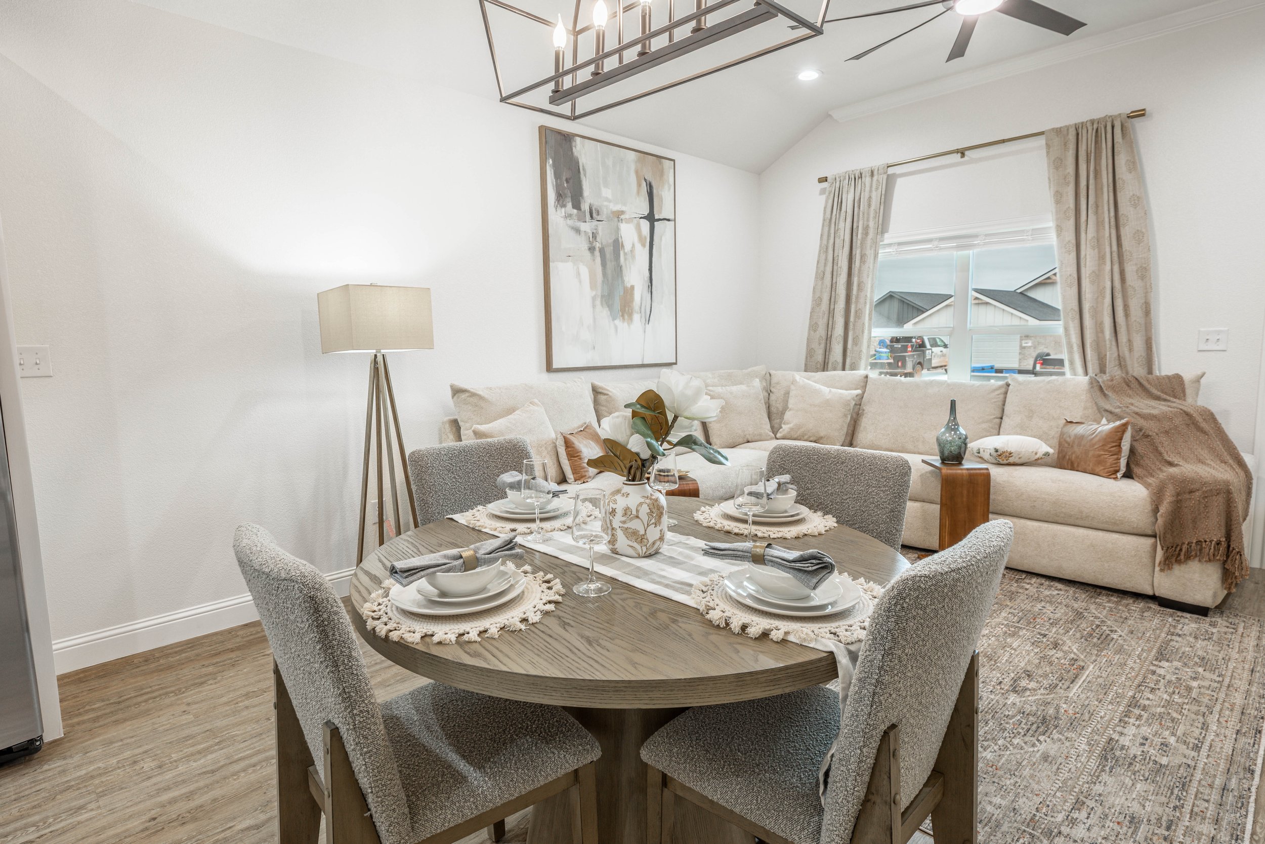 A living room with a wooden dining table set for a meal, surrounded by six gray upholstered chairs. There is a beige sectional sofa with pillows and a blanket. A large abstract painting hangs on the wall. A window with curtains shows parked cars outs
