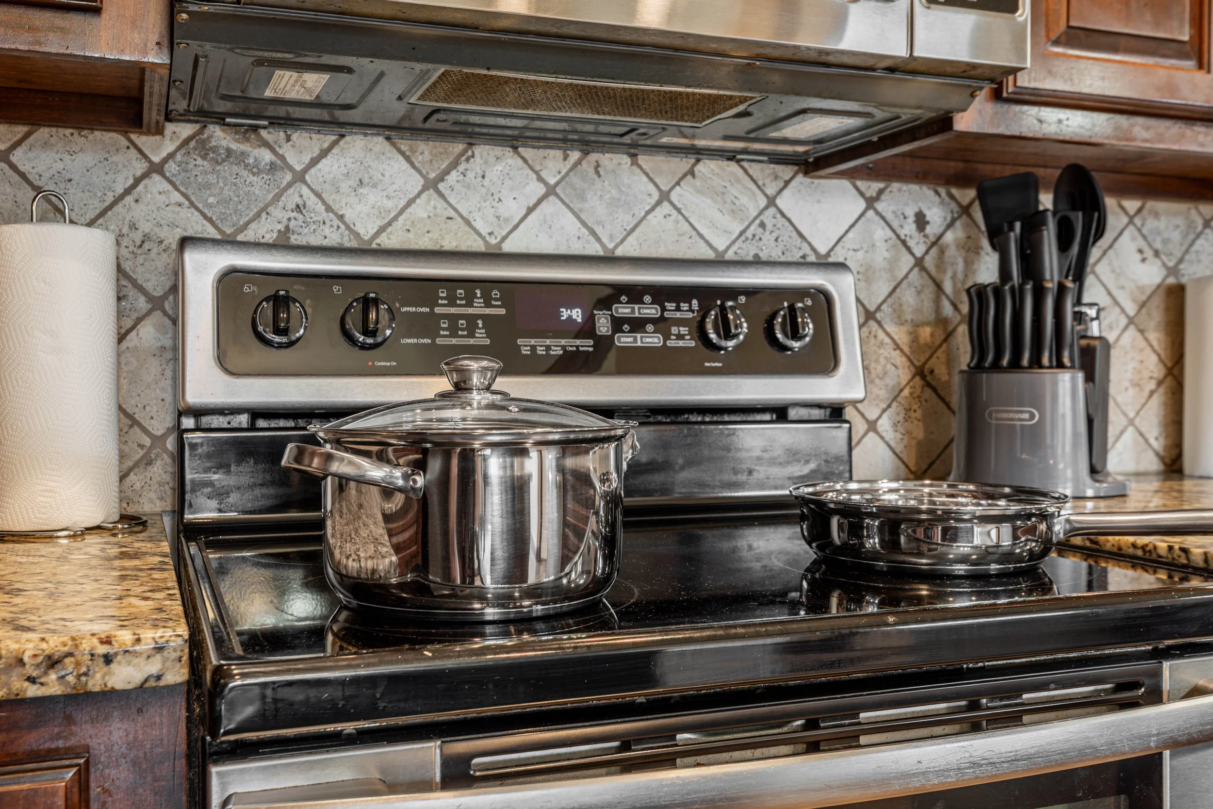 A kitchen stove with two pots on the burners, a paper towel roll on the left, a knife set in a holder on the right, and a tiled backsplash.