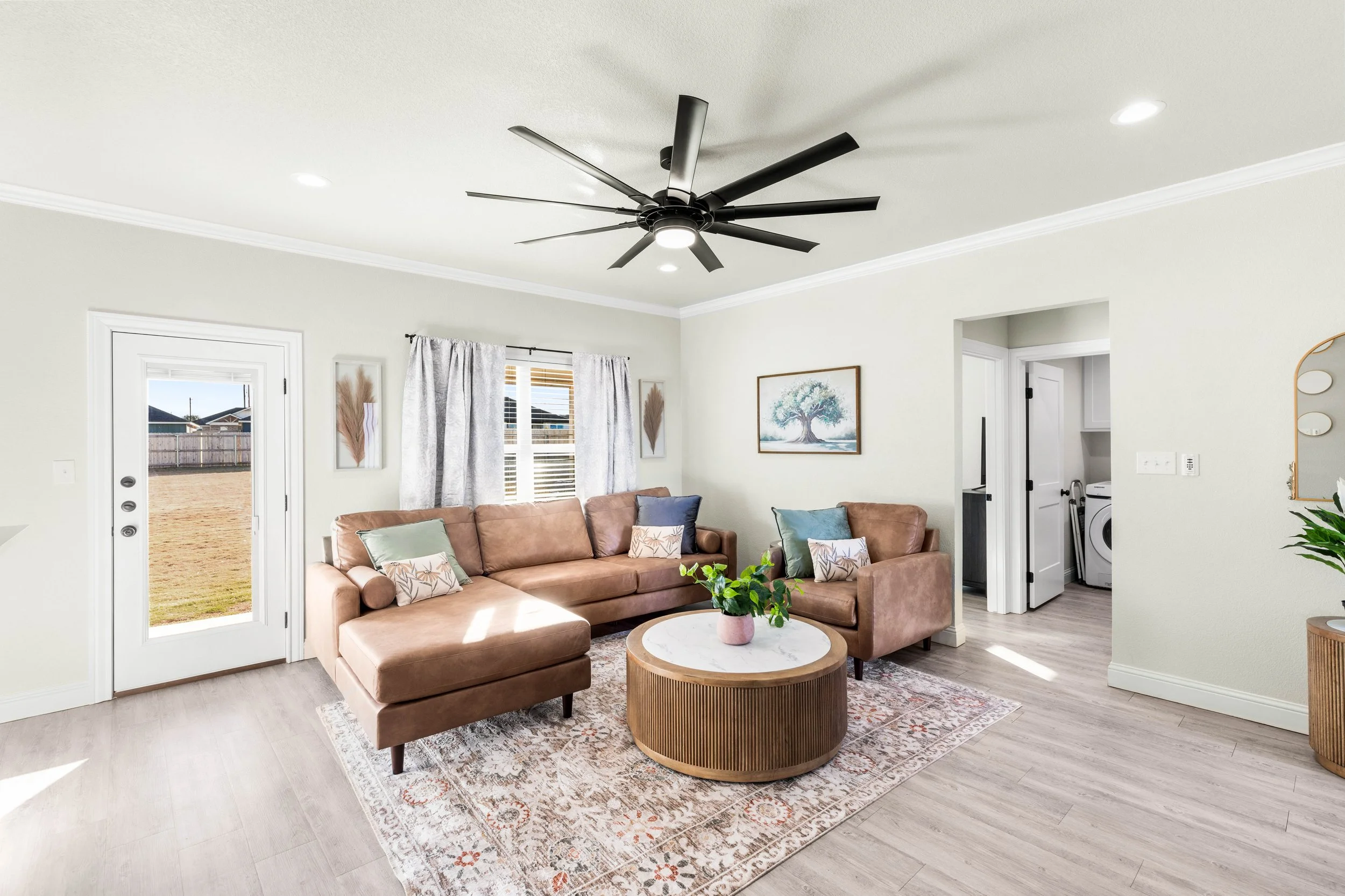 Living room with beige sectional sofa, white curtains, framed artwork of a tree, black ceiling fan, mirror, plant, and views of a fenced backyard through glass door.