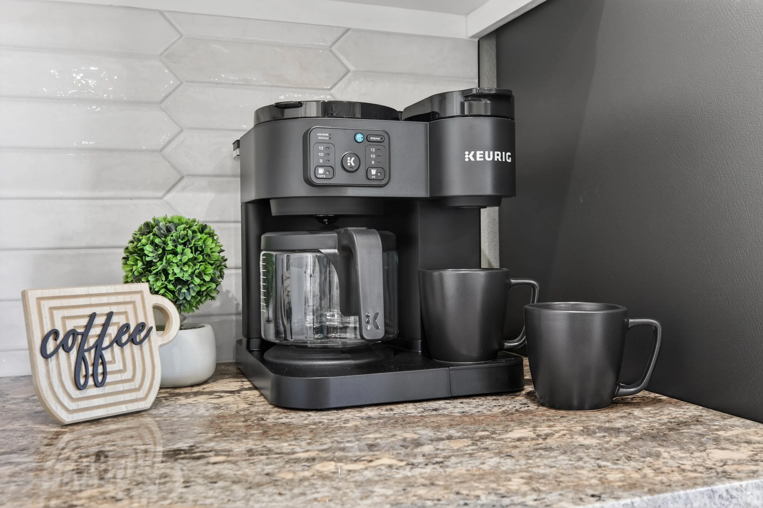 A Keurig coffee maker on a kitchen countertop next to a small green potted plant and a decorative sign that says 'coffee'. There are two black coffee mugs placed near the machine, and a glass coffee pot with a black handle is inserted into the machin