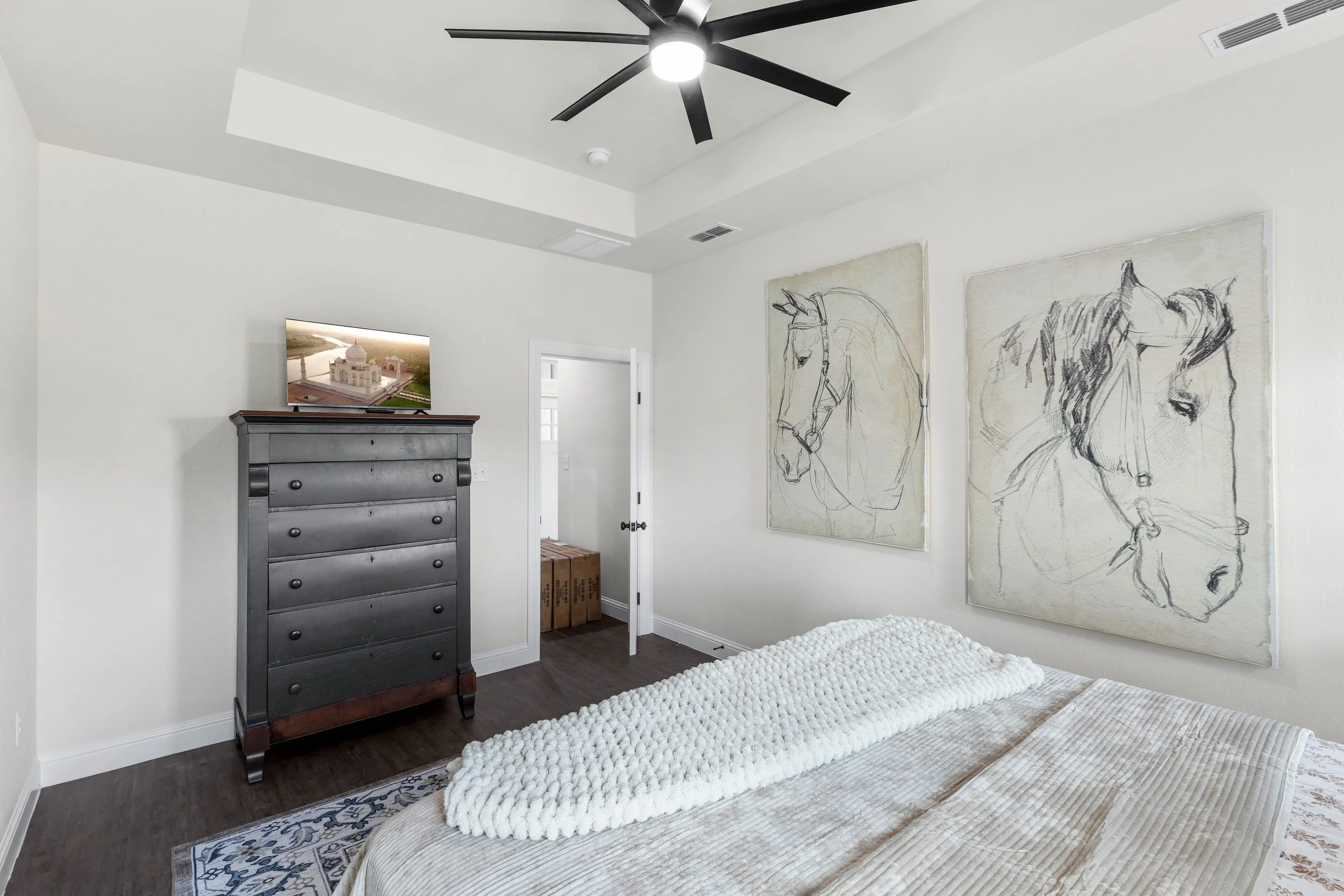 A bedroom with white walls and dark hardwood floors. There is a dark gray dresser with a flat-screen TV on top, displaying an image of a white church. Artwork of horse sketches hangs on the walls. A white textured blanket is draped over a bed in the 