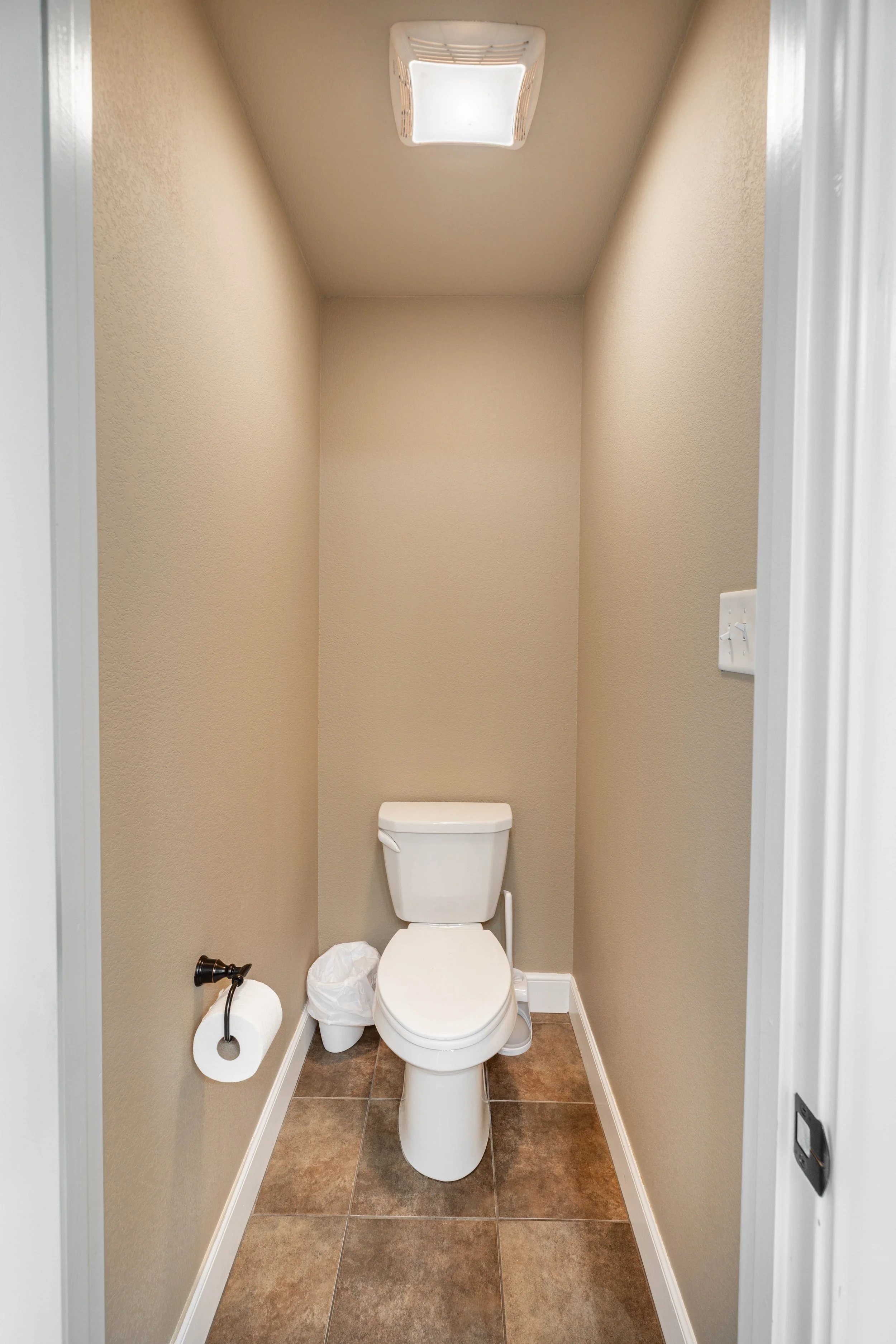 A small, narrow bathroom with beige walls and brown tiled floor. A white toilet is centered in the image, with a white toilet paper roll on a holder on the left wall, and a small trash can with a plastic bag on the floor. There is a window or vent hi