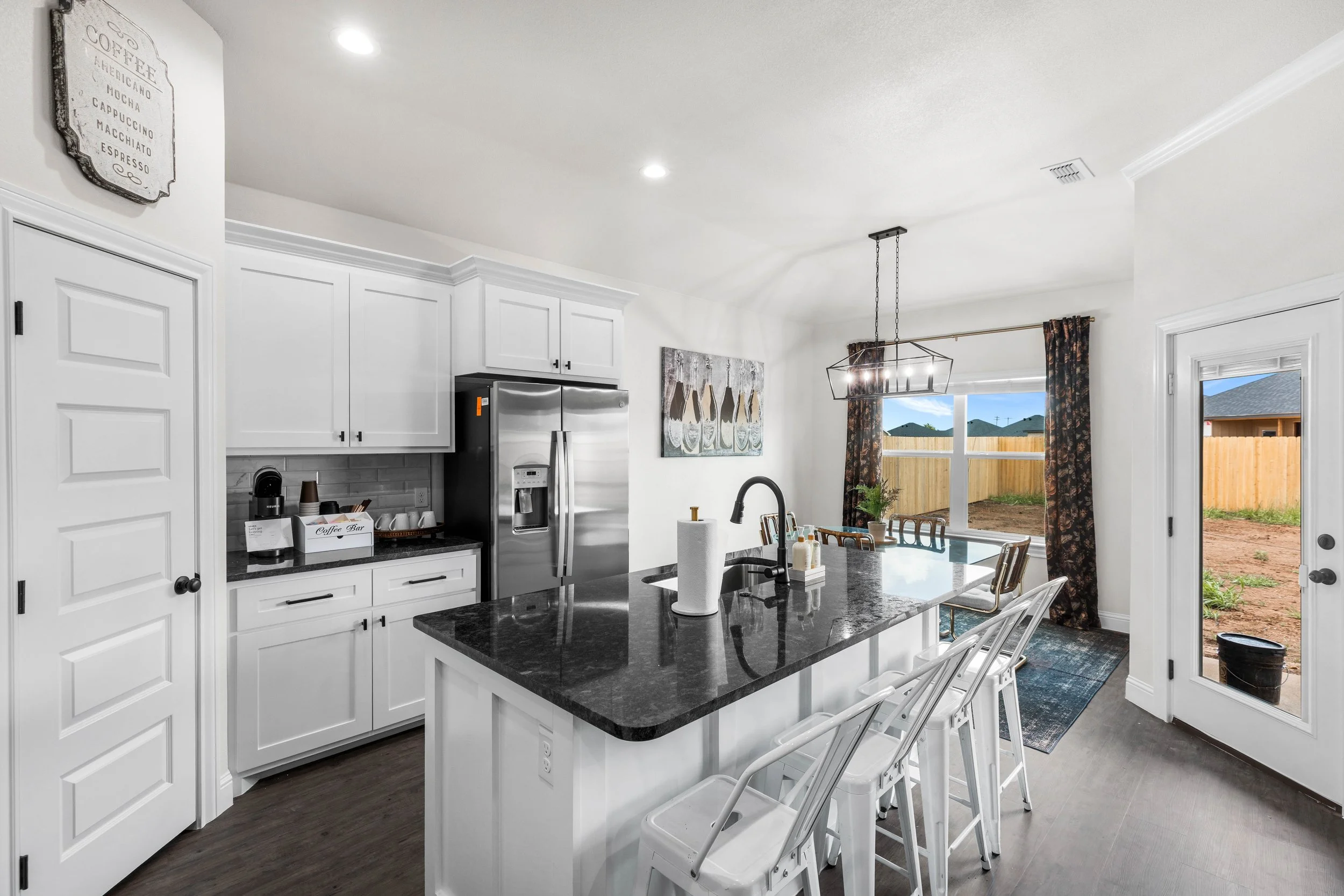 The image shows a modern kitchen with white cabinets, a black granite island, stainless steel refrigerator, and a window with floral curtains. There's a dining table with chairs, a painting on the wall, and a chandelier hanging from the ceiling. Outs