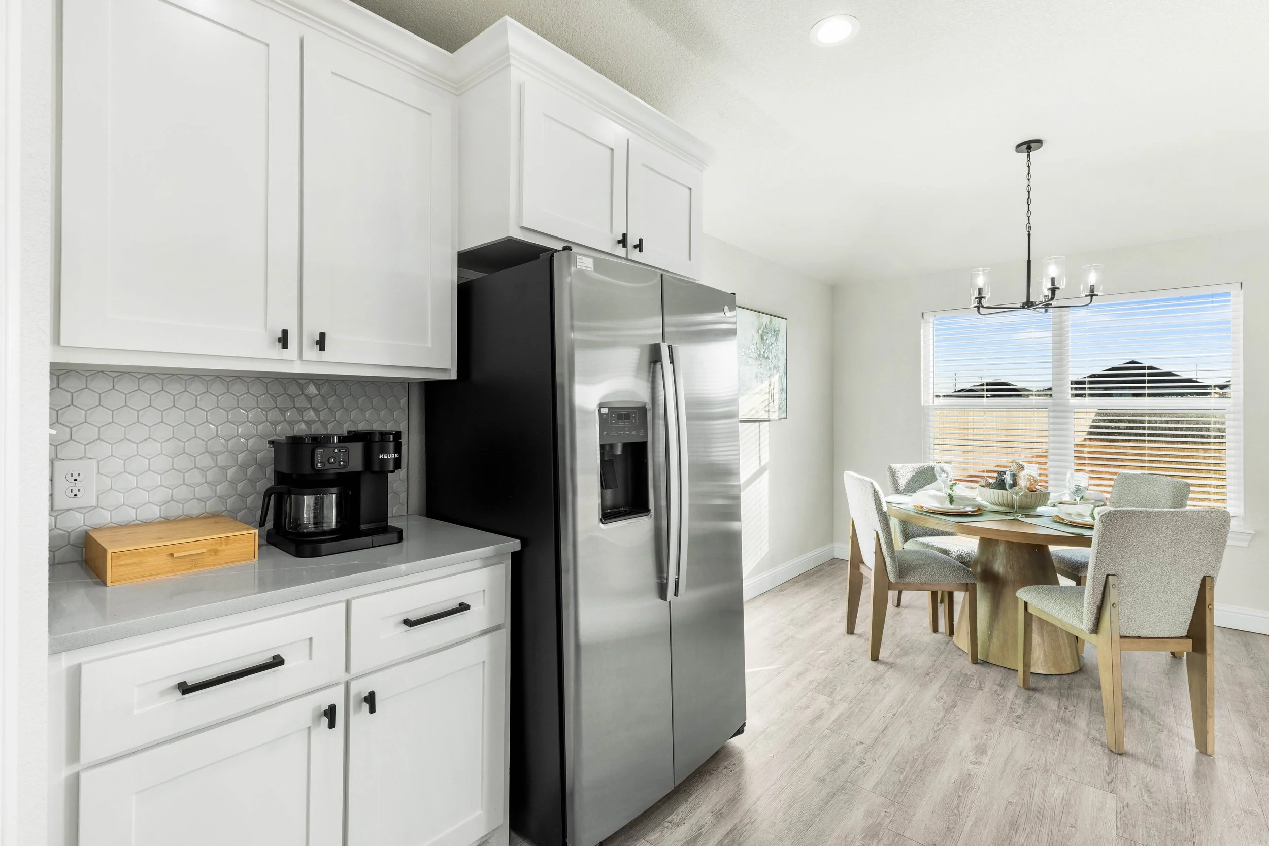 Kitchen with white cabinets, a stainless steel refrigerator, a black coffee maker on the countertop, a wooden bread box, and a dining area with a round wooden table surrounded by four upholstered chairs, adjacent to windows with blinds.
