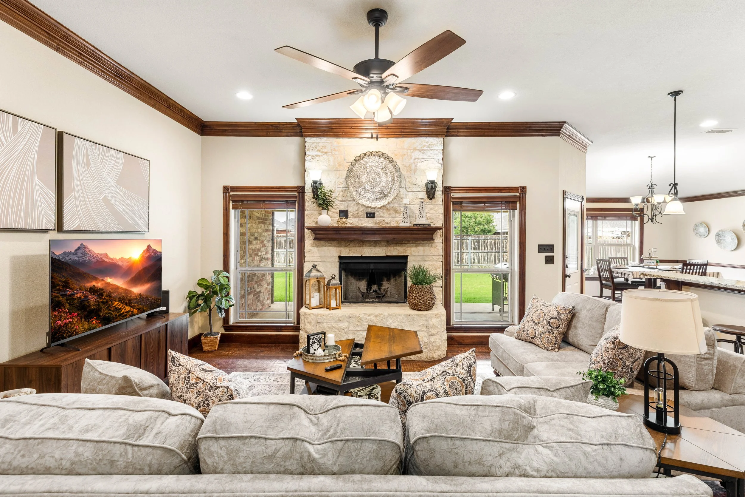 A cozy living room with a stone fireplace flanked by two large windows, wooden crown molding, and a ceiling fan with light fixtures. The room features a sectional sofa with patterned pillows, a wooden coffee table, and a TV displaying a mountain land