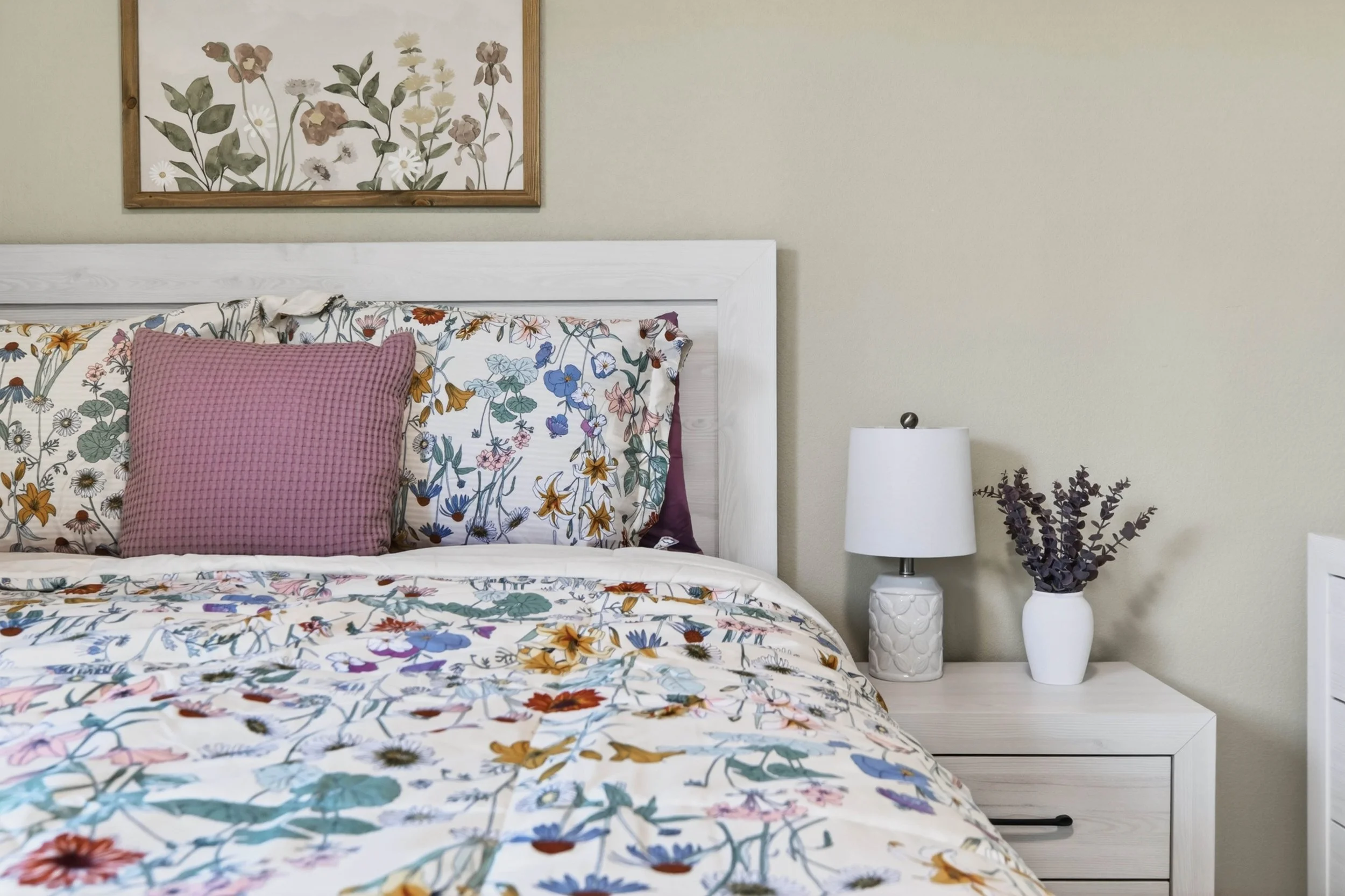 A bedroom with a wooden framed bed, floral bedding, a pink textured pillow, a white nightstand, a white lampshade, and a vase with purple flowers, with a framed floral art on the wall.