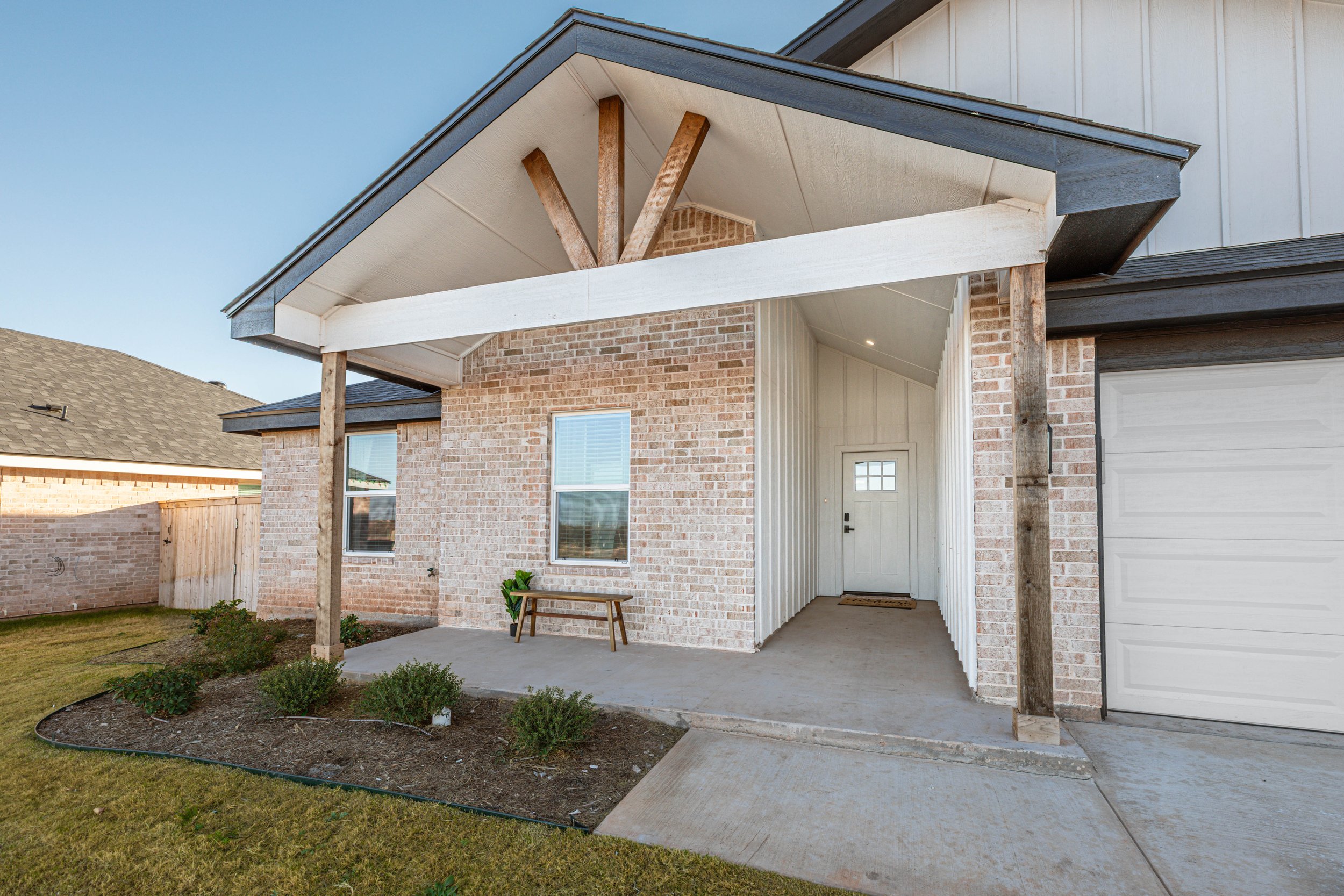 Back porch and entryway of a brick house with a small yard and garden beds, featuring a white door, two windows, and a covered patio area supported by wooden beams.