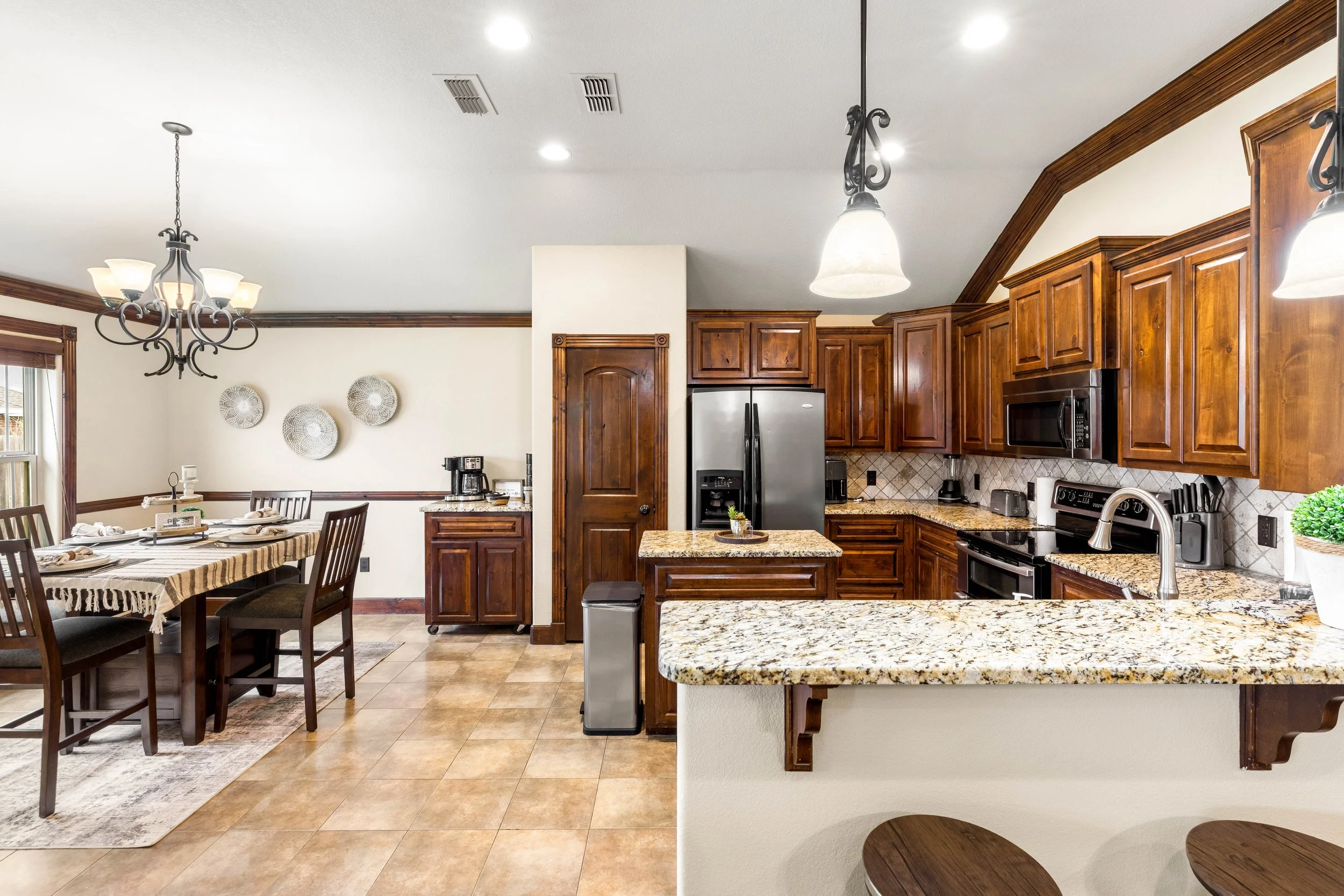 A kitchen with wooden cabinets, a granite countertop, and stainless steel appliances. There is a breakfast bar with two wooden stools, and a dining area with a table and chairs decorated with a striped tablecloth. A chandelier hangs over the dining t