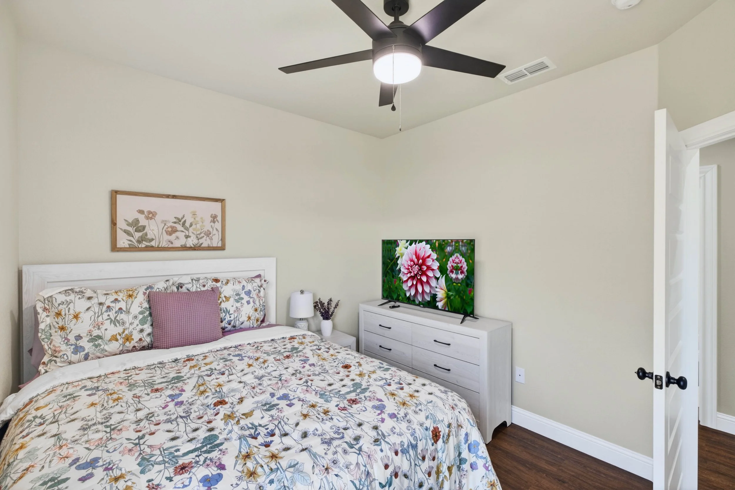 A bedroom with a floral bedspread and pillows, a white dresser with a TV showing a flower, a nightstand with a lamp, wall art, ceiling fan, and a partly open door.