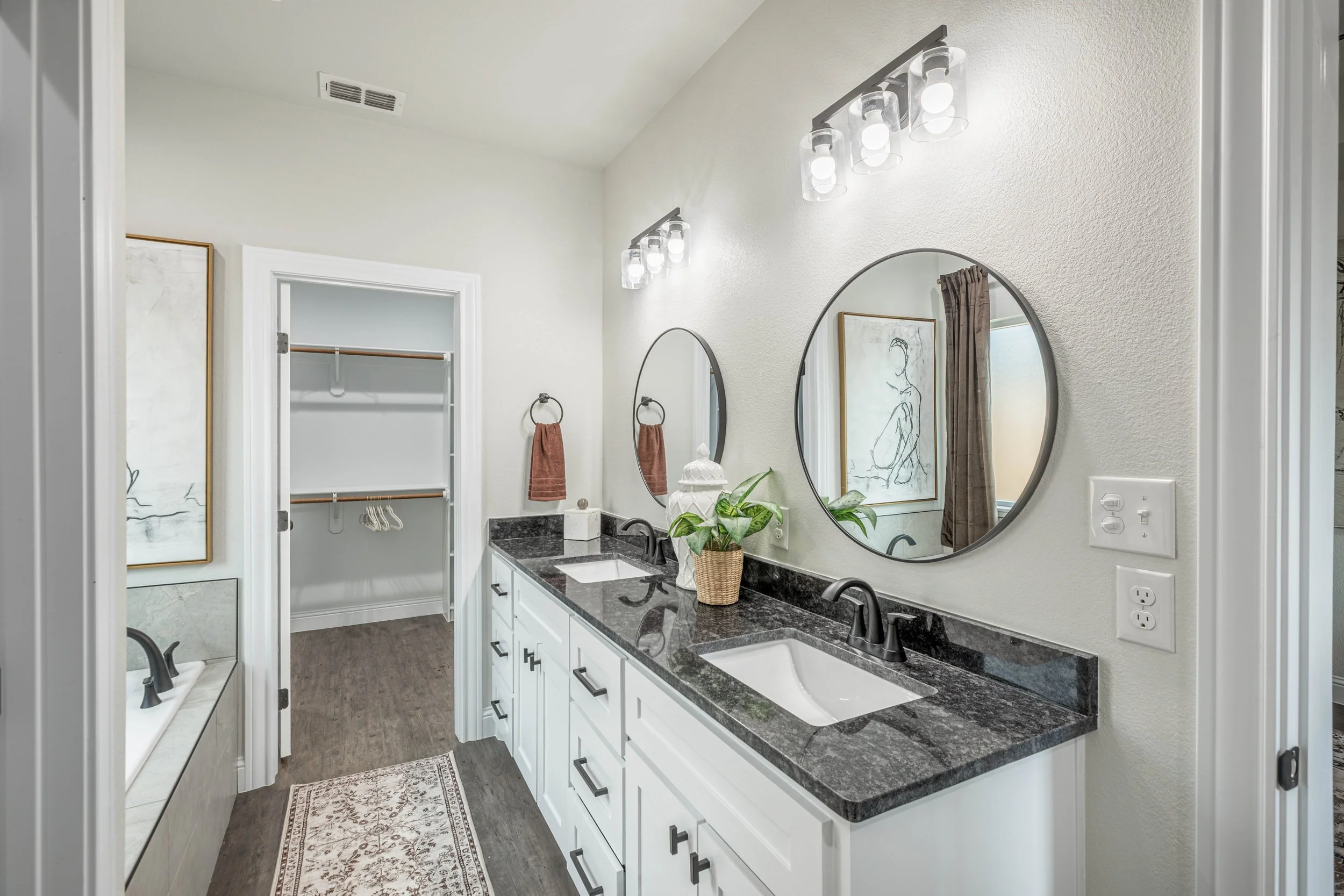 A modern bathroom with a white double vanity, black granite countertop, two oval mirrors, and a small potted plant. There are two wall-mounted light fixtures above the mirrors, a framed abstract art piece, a towel ring with a brown towel, and a parti