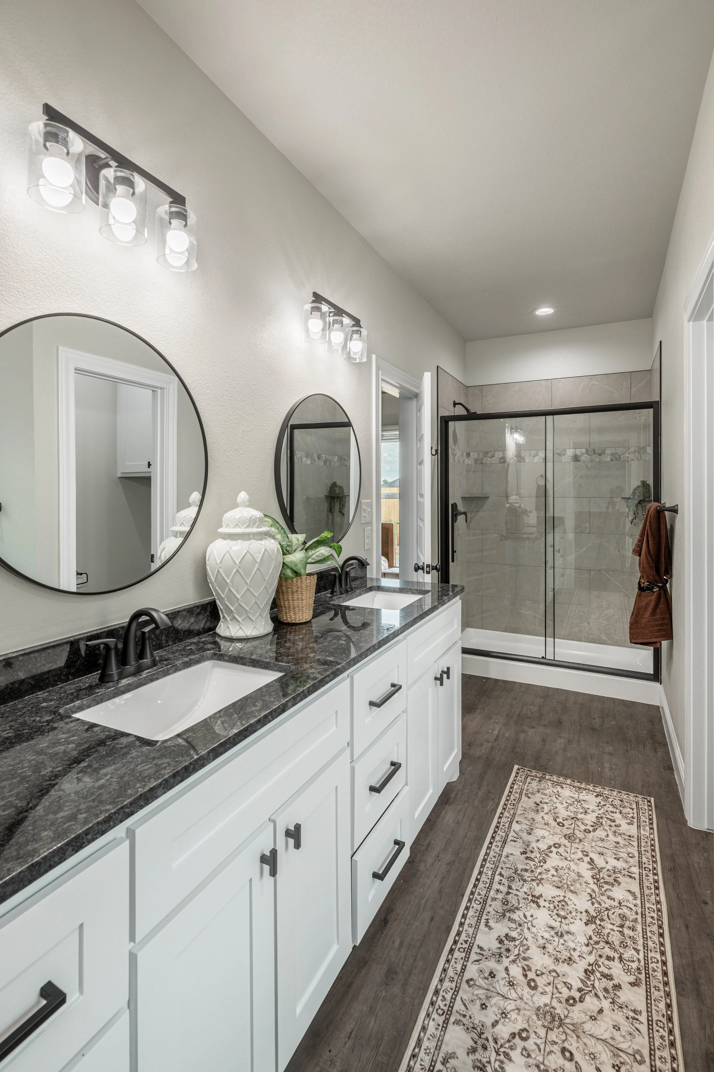 Bathroom with dual sinks, round mirrors, decorative vases, plants, shower with glass door, and a brown towel on a towel rack.