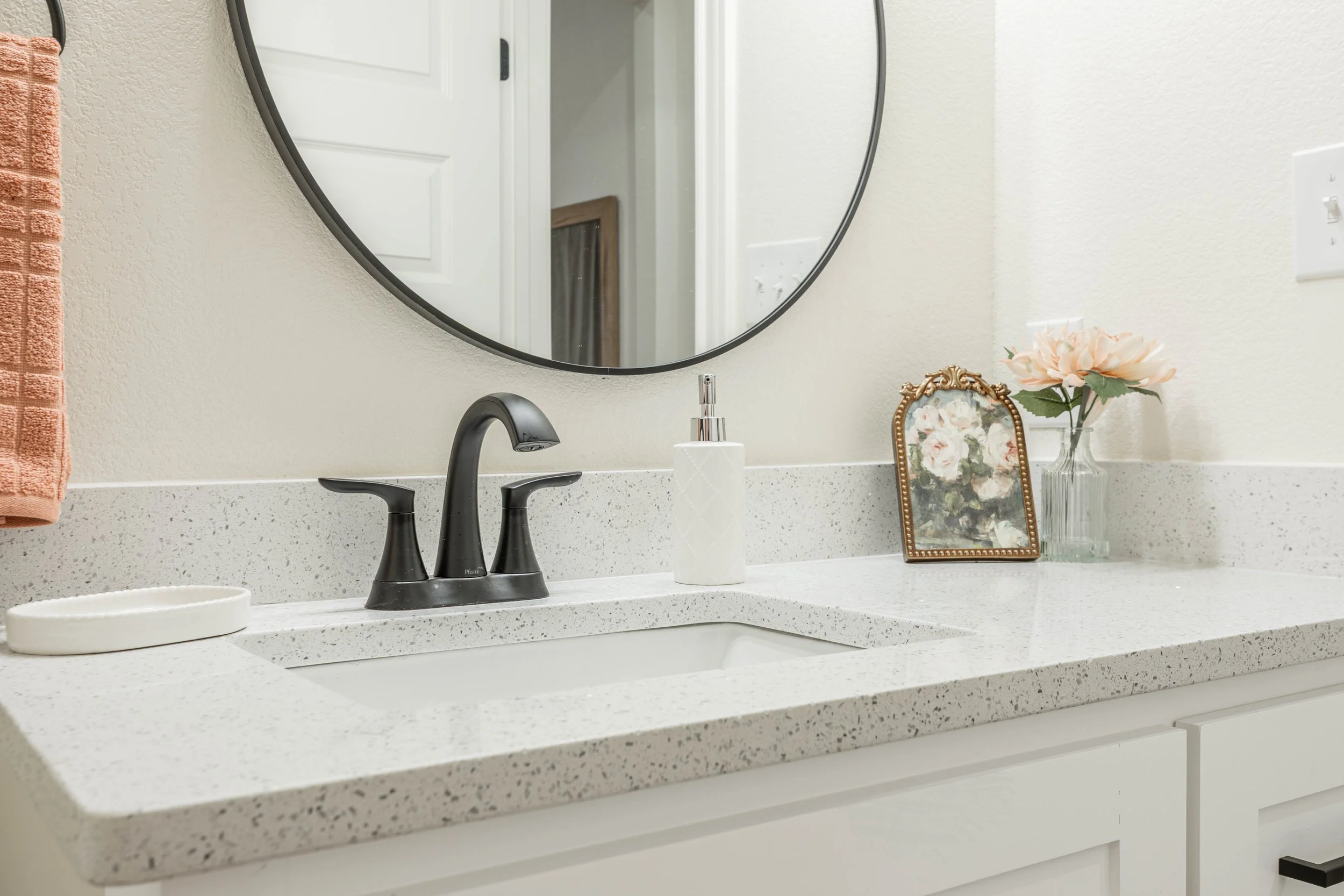 Bathroom countertop with a white speckled surface, black faucet, a soap dispenser, a decorative picture frame with floral painting, pink artificial flowers in a glass vase, and a white dish.