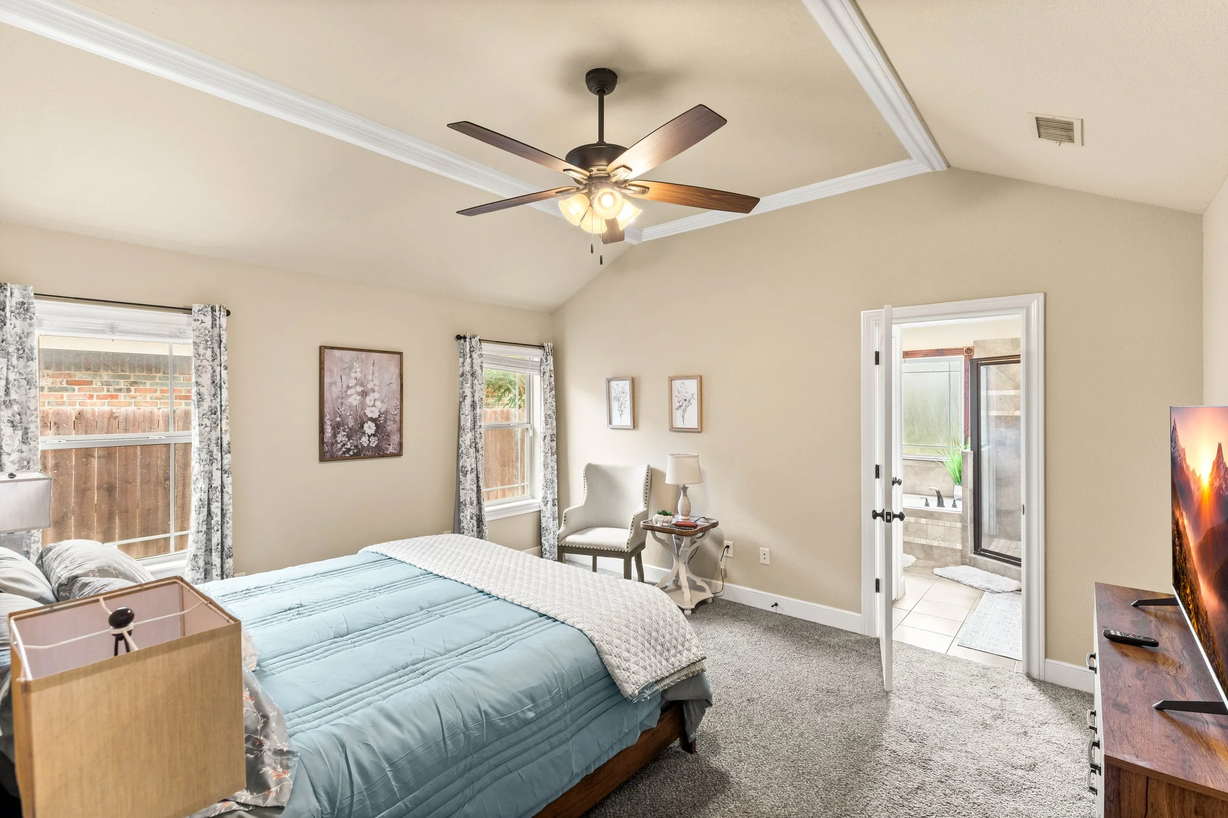 A bedroom with beige walls, a ceiling fan with lights, two windows with floral curtains, a white armchair, and a door leading to a bathroom with a bathtub.