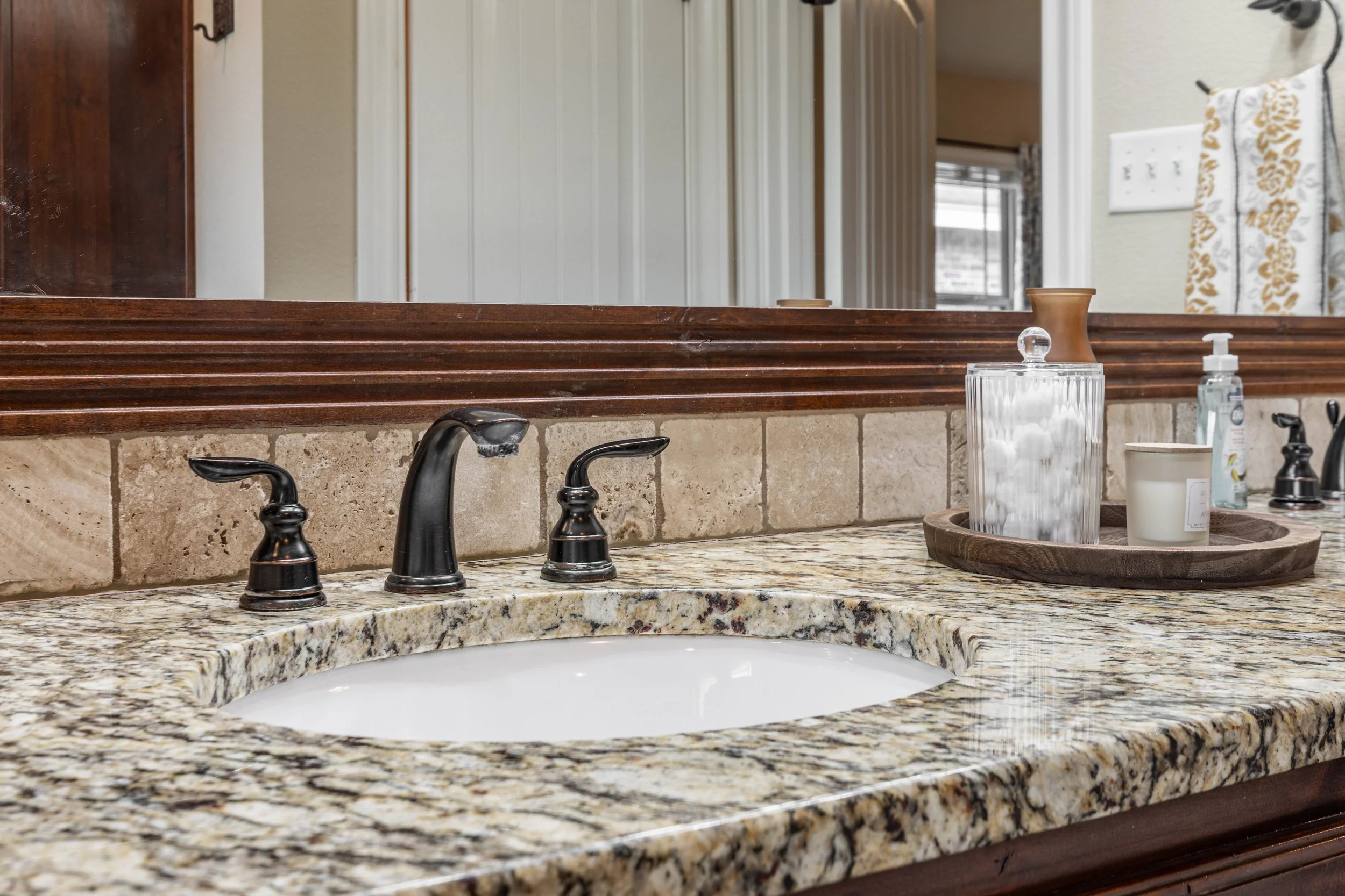 Bathroom vanity with a granite countertop, a built-in oval sink, black faucet fixtures, a wooden tray holding a clear glass container with cotton balls, a small container, and a soap dispenser. There is a large mirror above the sink, and part of a pa