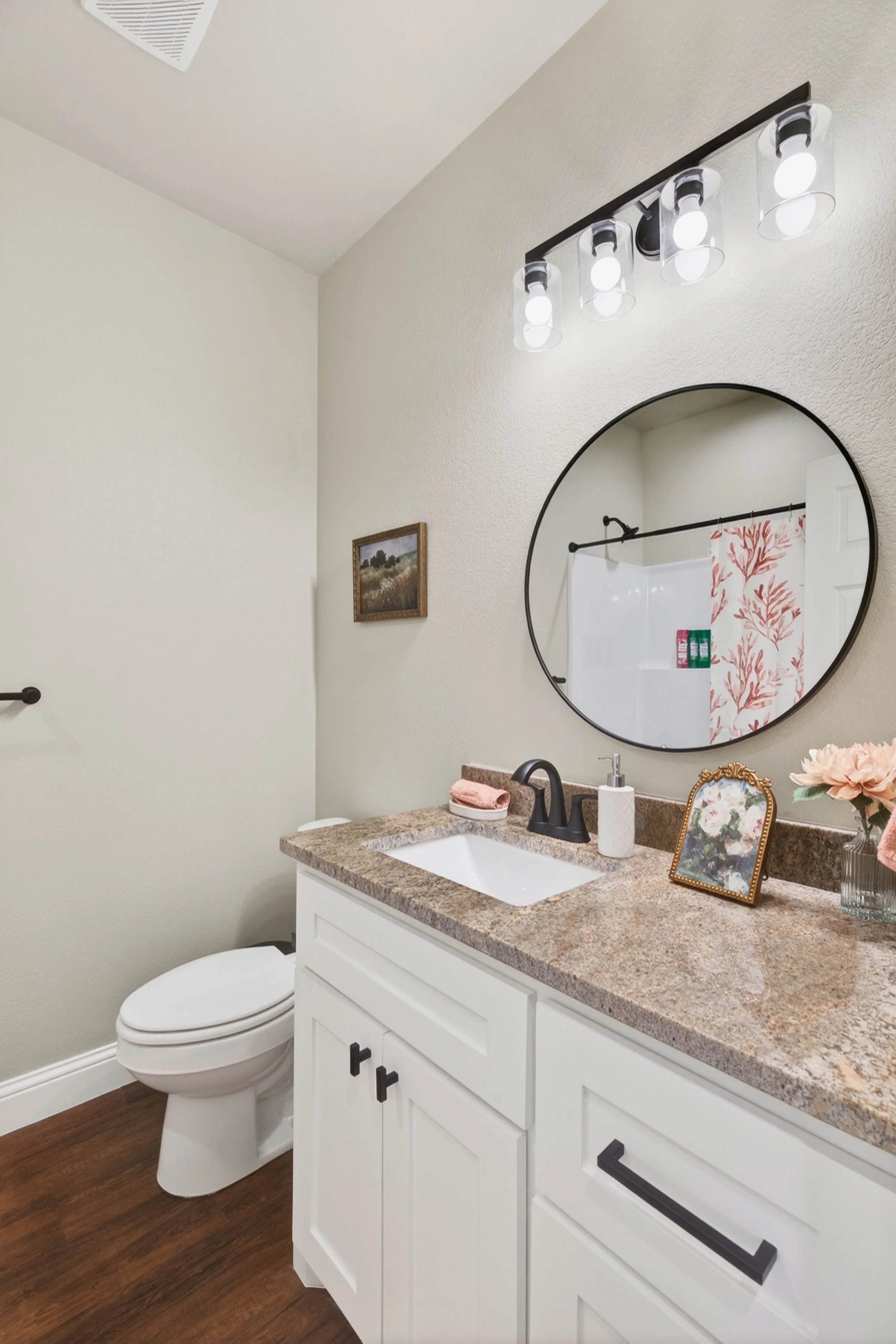 Bathroom with white vanity and granite countertop, round mirror, black faucet, framed floral picture, vase with pink flowers, soap dispenser, and shower curtain with coral pattern; toilet to the left, wall decoration and lighting fixture above mirror