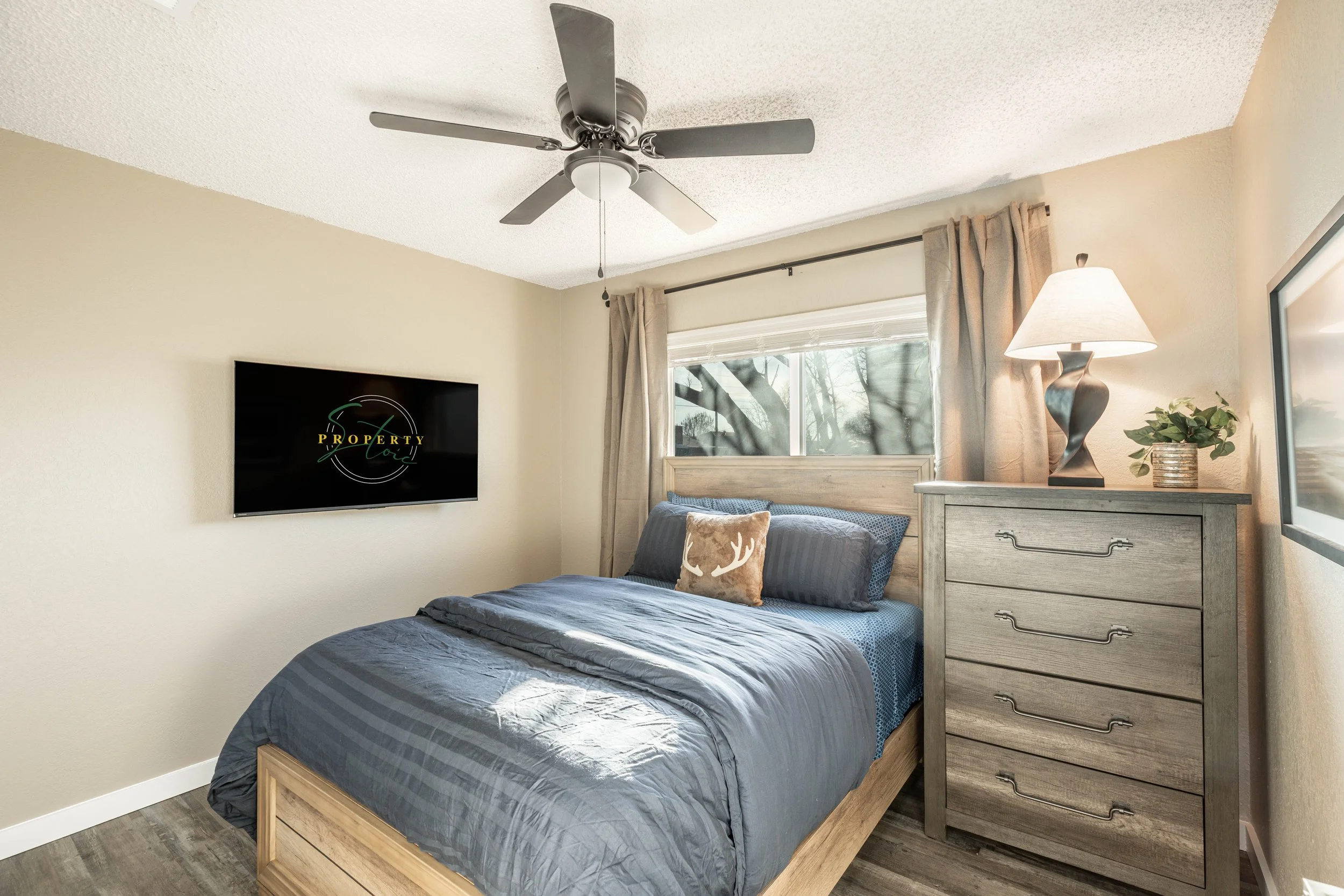 A bedroom with a wooden bed frame, gray bedding, a decorative pillow, a nightstand with a desk lamp and a small plant, a window with beige curtains, a wall-mounted TV, and a ceiling fan.